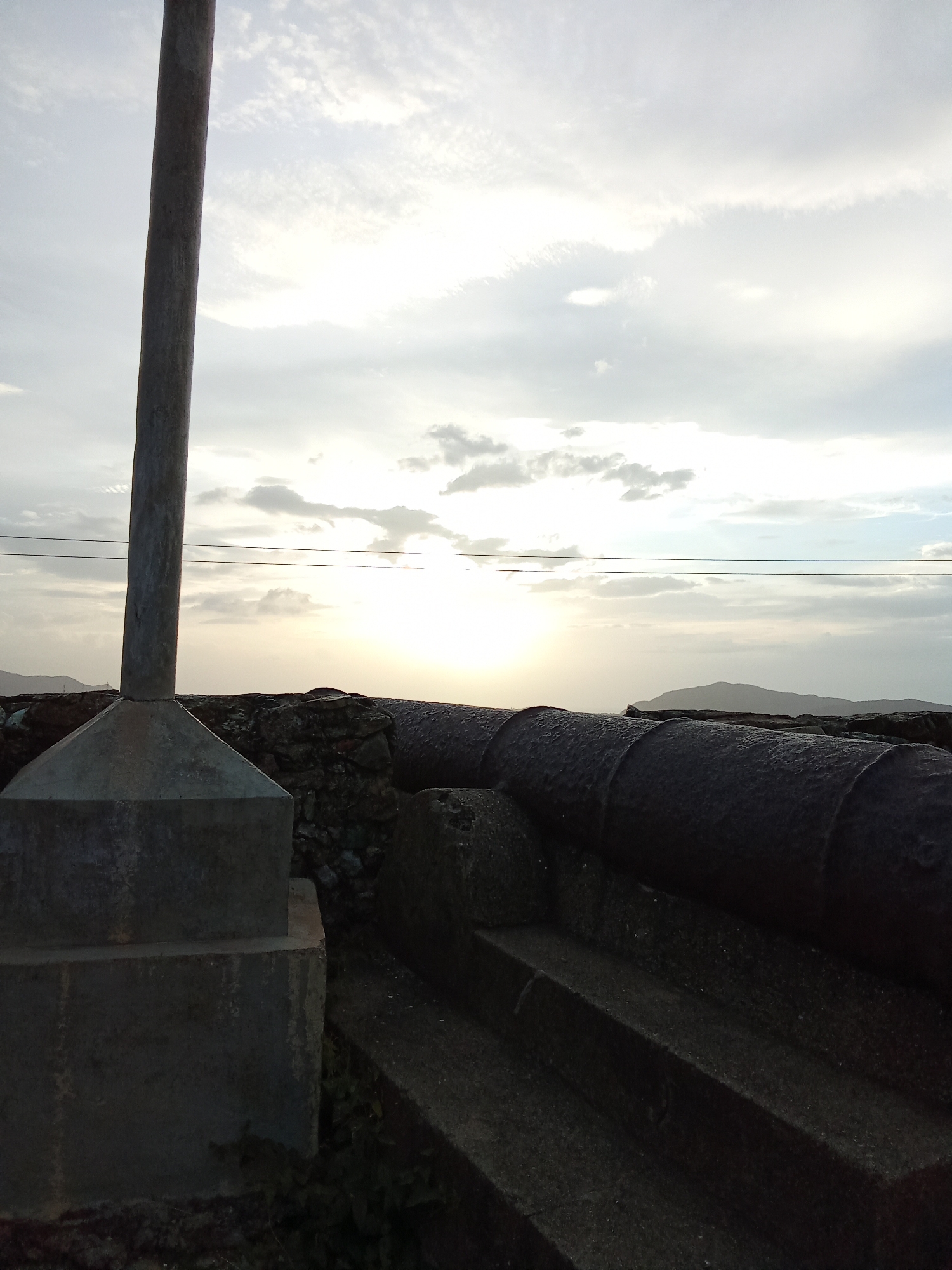 A low-angle shot depicts a historic scene, likely a fort or historical site. A large, dark cannon rests on a stone platform with stepped levels leading up to it. The cannon points towards the horizon where the sun is setting or rising, casting a bright glow through the clouds. A concrete pillar or post stands to the left, partially obscuring the view. In the background, a coastline or distant hills are faintly visible. Electrical wires stretch across the upper part of the frame, adding a modern element to the old setting. The location is identified as being in the city of Santa Ana, Venezuela. No people are visible.