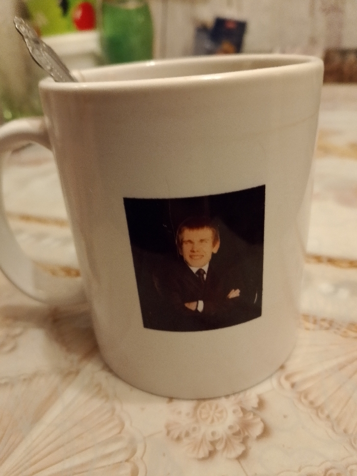 A white mug sits on a beige patterned surface. A silver spoon is visible inside the mug. A black square sticker adorns the mug, displaying a portrait of a man in a suit with his arms crossed. The background shows a blurry kitchen setting with green and red items.