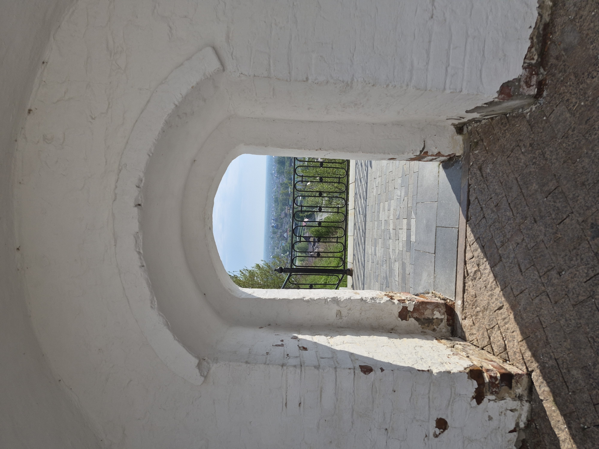 A high-angle shot captures a view through a white, arch-shaped opening in a brick structure in Tobolsk, Russia. The opening leads to a view of a cityscape with greenery, a black metal railing, and a paved area. The arch is constructed from white-painted bricks, showing signs of wear and chipping. To the right of the opening, the structure transitions into a darker, textured brick. The exterior environment includes trees, rooftops, and a distant skyline under a light blue sky.