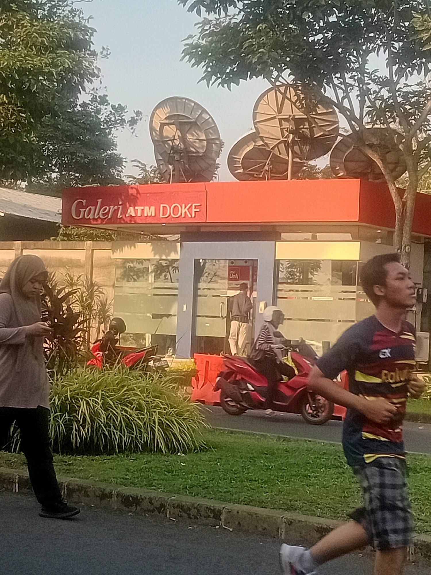 In Bacin, Indonesia, a group of people are seen near a red building with satellite dishes on its roof. A woman wearing a grey hijab and black pants is walking while looking at her phone. A man in a blue and red shirt with yellow stripes and plaid shorts is running. A red motorcycle is parked near the building, with two individuals on it, one wearing a white helmet. The building has a sign that reads "Galeri ATM DOKF". The area appears to be an outdoor setting with trees and grass.