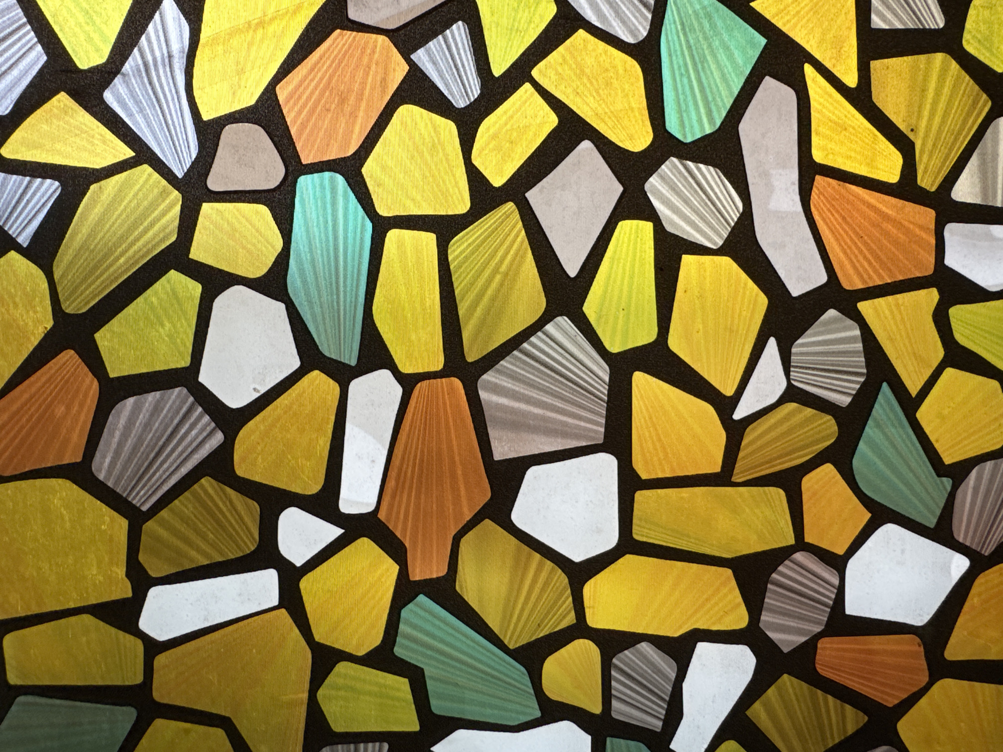 The image shows a close-up view of what appears to be a decorative window or panel. The design features irregularly shaped pieces of colored glass or translucent material, resembling a mosaic or stained glass pattern. The pieces are primarily yellow, orange, blue-green, white, and gray, separated by a dark brown or black grid. The pieces feature textures that look like converging stripes, and have varying levels of opacity. The image does not contain any people or other objects other than the panel. The location of the image is not possible to determine from the visual elements.