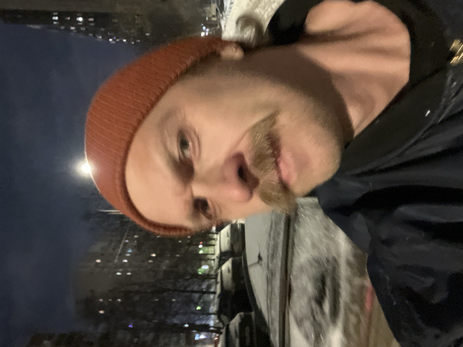 A close-up selfie of a man with a mustache and an orange beanie. He is wearing a dark jacket. The background appears to be an urban nighttime scene, with snow on the ground and buildings visible in the distance. The image is rotated, making it appear sideways. The man's face is the primary focus, taking up most of the frame.
