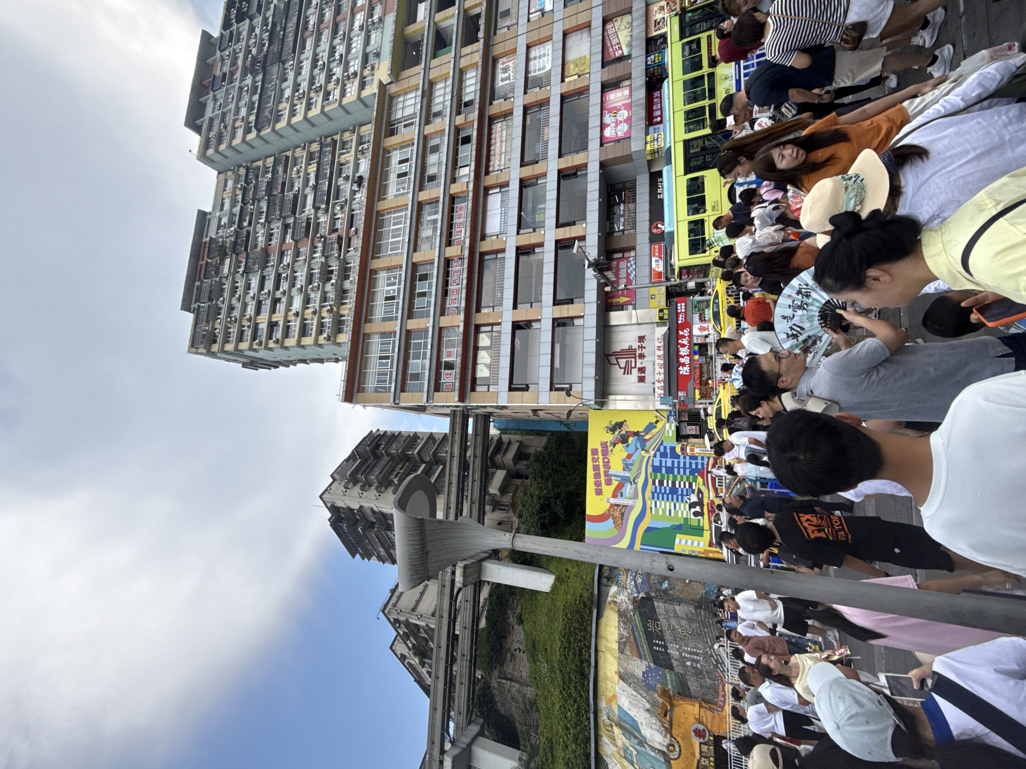 The image captures a bustling street scene in Yuzhong District, China, with towering apartment buildings dominating the background. A monorail track curves above a lush green hillside on the left. In the foreground, a crowd of people navigates a busy pedestrian area. Several individuals are visible holding cell phones, and one person is fanning themselves with a large, decorated fan. A bright yellow bus is parked on the street, with advertisements displayed on its side. Vibrant murals adorn a wall on the left side of the frame, adding a splash of color to the urban landscape. The sky above is partly cloudy.