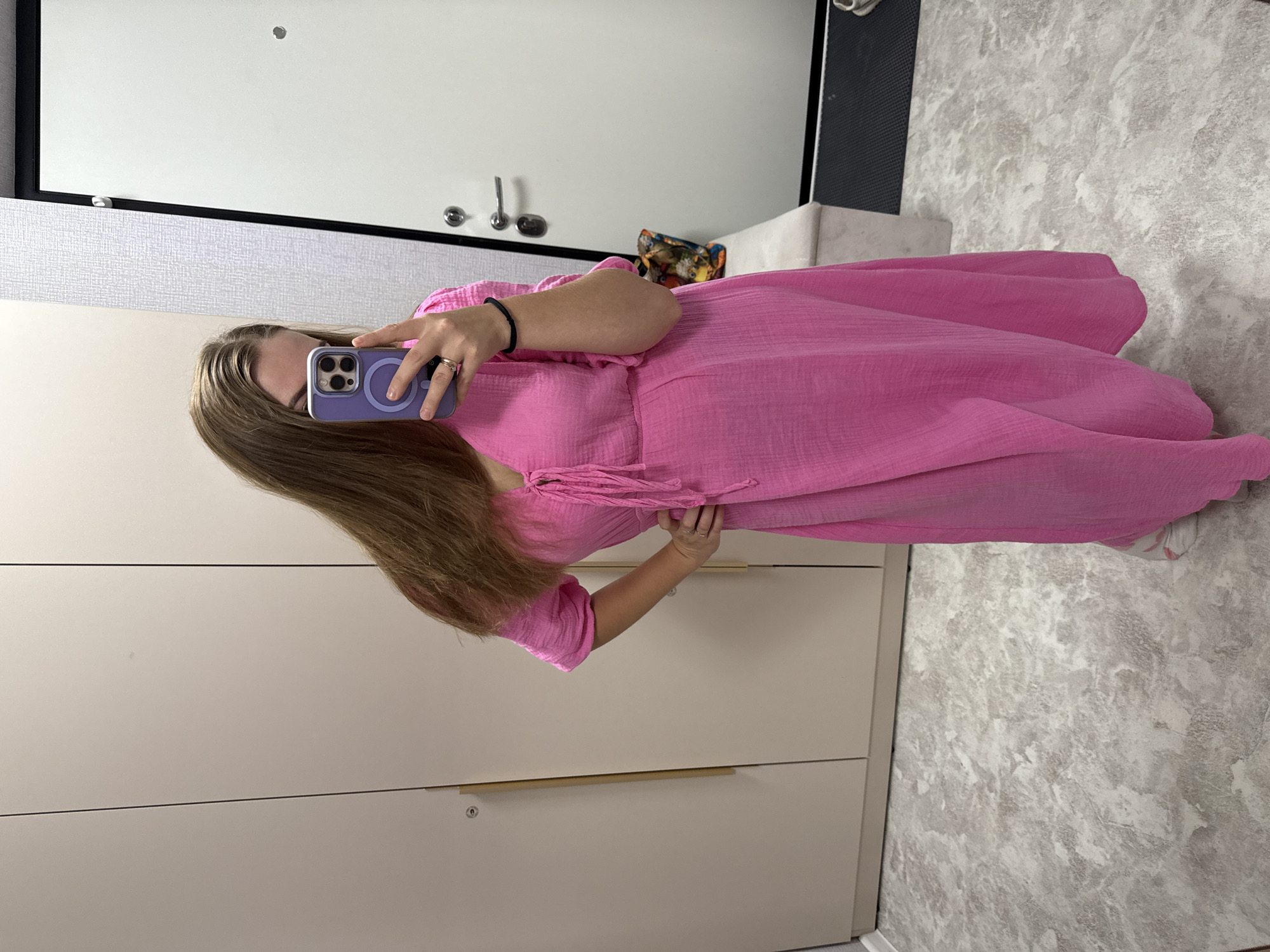 In a dimly lit room, a person stands in front of a mirror, taking a selfie. They are wearing a bright pink dress and holding a smartphone in their left hand. The room appears to be a dressing room or hallway, with a large wardrobe on the left and a white door on the right. The floor is covered in a grey, marbled tile. The lighting in the room is soft, creating a muted atmosphere. The image does not provide enough information to determine the location in Kazan, Russia.