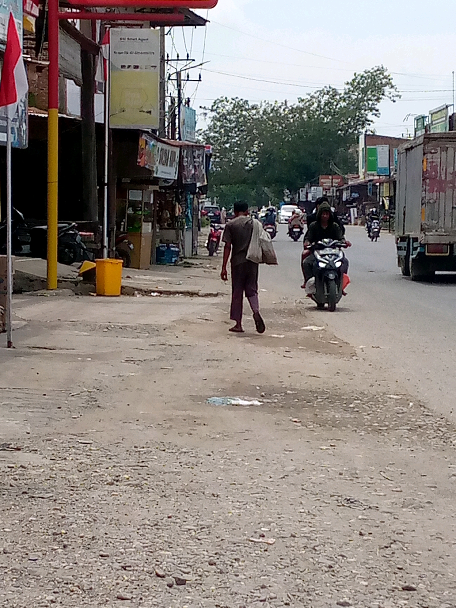 The image depicts a street scene in Keudebayu, Indonesia, under an overcast sky. In the foreground, a man walks away from the camera, carrying a large sack over his left shoulder. He is wearing dark pants and a light-colored shirt. To his left, a yellow trash bin is visible on a gravelly surface.
The midground shows a road with several motorcycles. One motorcycle, with two riders, is prominently featured, with the driver wearing a hooded jacket. Other motorcycles are visible further down the road, with drivers and passengers. A white car is also present in the distance.
The background features buildings lining the street, some with signage. A large tree with dense foliage is situated behind the buildings. Power lines are strung across the sky. On the right side of the image, a portion of a large truck is visible, indicating its presence on the road. The overall environment appears to be a commercial or residential area along a street.
