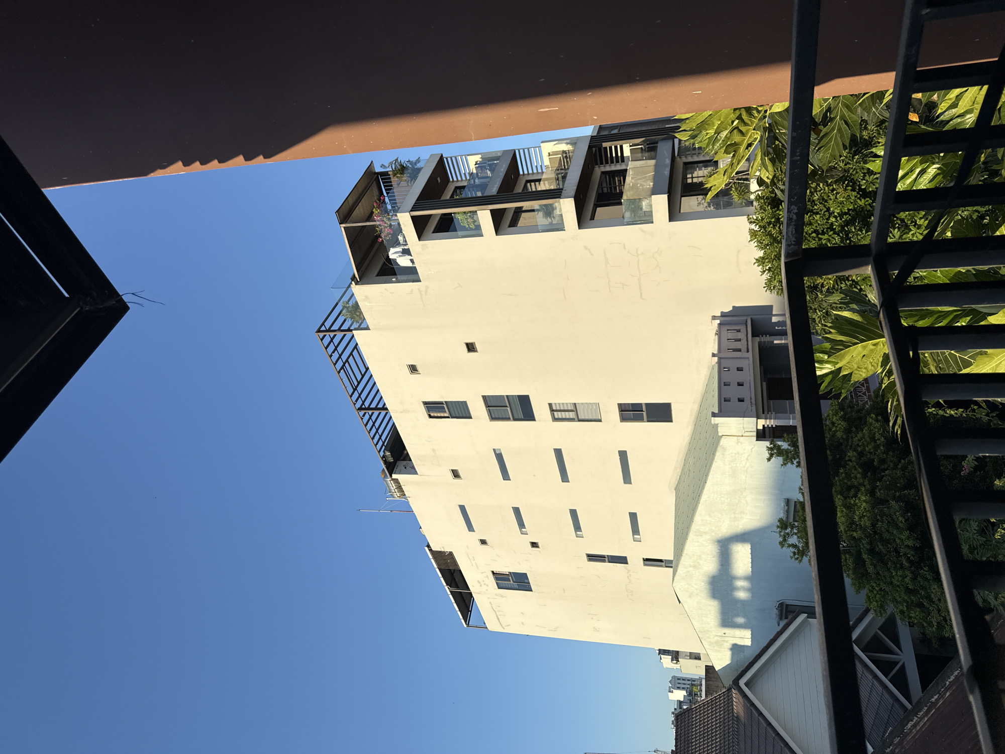 A low-angle shot shows a multi-story building in Đà Nẵng, Vietnam, with a pale exterior, numerous windows, and rooftop structures. Partial views of roofing and a metal staircase obscure the foreground. Clear blue sky fills the space around the building. Nearby trees and neighboring buildings are visible at the base of the main structure.