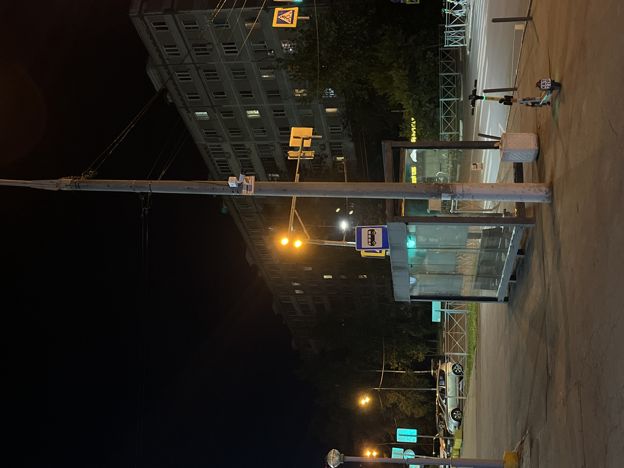 At night in Mytishchi, Russia, a street scene unfolds. A bus stop shelter, constructed with glass and metal, stands prominently in the foreground. The shelter features a blue and white bus stop sign with a pictogram of a bus. Adjacent to the shelter, a tall utility pole supports electrical wires and a security camera. On the sidewalk near the bus stop, a white scooter with a license plate reading "KA1 27P" is parked.
Across the street, a multi-story apartment building with numerous lit windows dominates the background. Traffic lights, one with two yellow lights, are visible. A pedestrian crossing sign, a yellow square with a walking figure, is mounted on a pole. The street is illuminated by streetlights and the lights from the building. Trees line the side of the road behind the bus stop. In the distance, cars are parked, and a chain-link fence separates the sidewalk from the street. The overall atmosphere is urban and nocturnal.