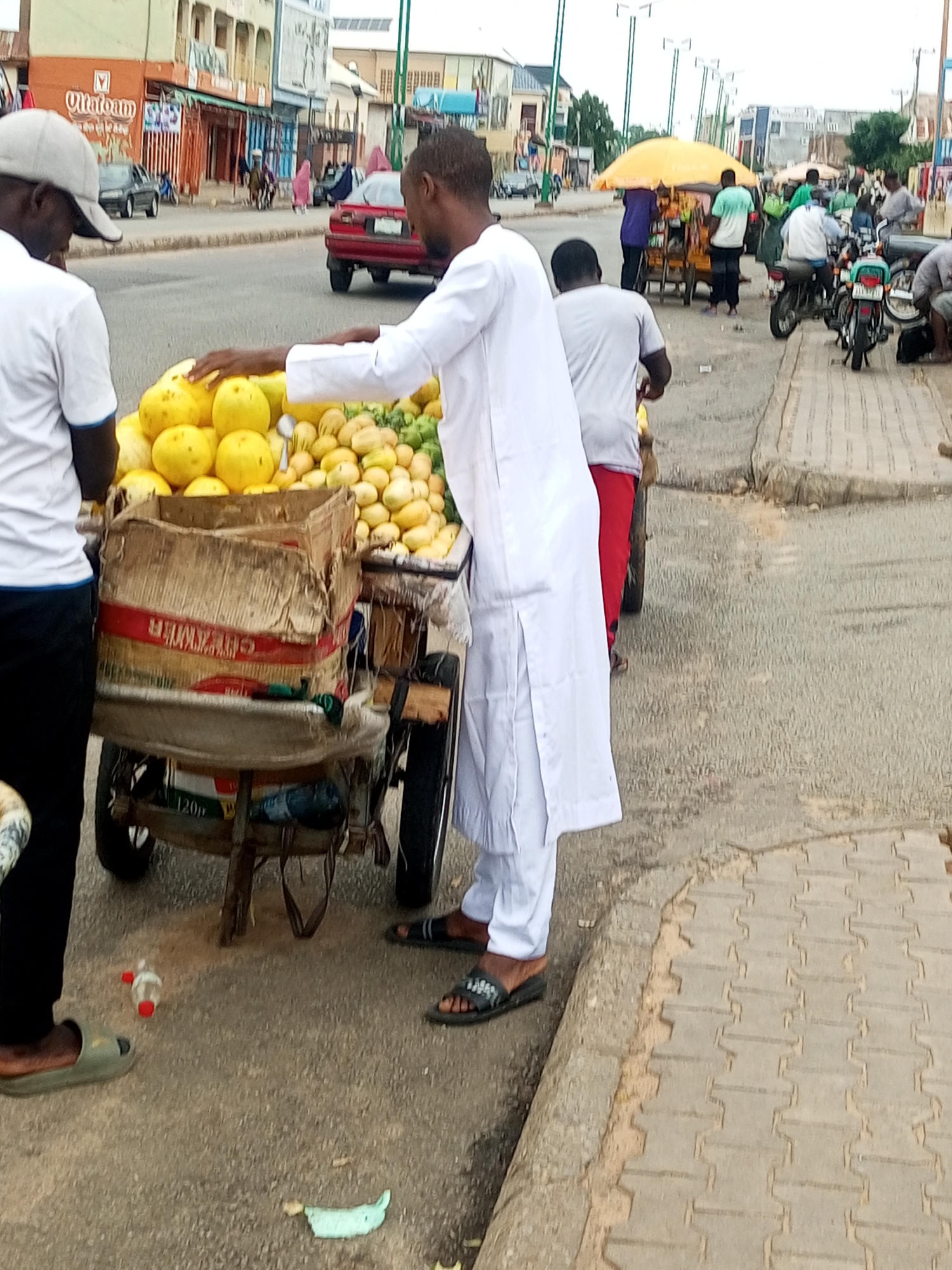 In Arawa, Nigeria, a street vendor is captured from the side view, standing next to a cart filled with yellow fruits. He wears a white, long-sleeved tunic and pants. Another man, partially visible on the left, wearing a white t-shirt and a grey baseball cap, appears to be interacting with the vendor. The cart, made of a damaged cardboard box with red and white banding, rests on a wheeled frame. Behind the vendor, other street vendors with a yellow umbrella and a food stall are visible, along with people on motorcycles and in vehicles on the street. The scene unfolds on a paved street next to a tiled sidewalk, with buildings lining the background.