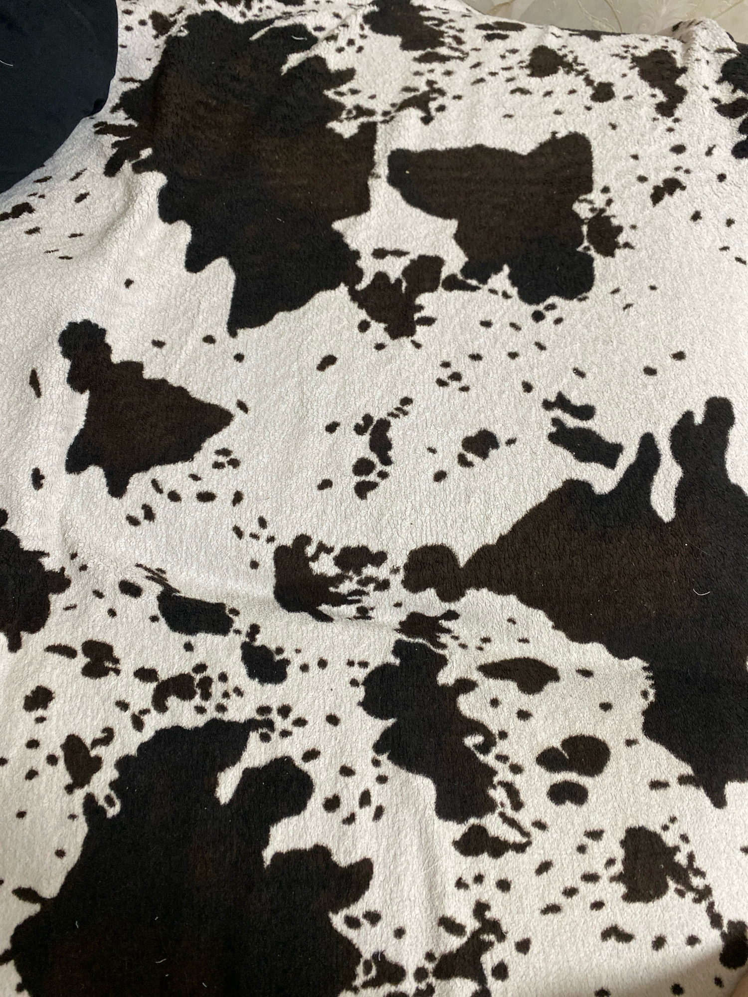 The image displays a close-up, overhead view of a cowhide-patterned blanket or rug. The pattern consists of irregular black splotches on a white background, with some areas featuring smaller black dots scattered across the white. The texture appears soft and somewhat fuzzy. A portion of a black garment is visible in the upper left corner of the image. The location of this item within Ivanteyevka, Russia, is not discernible from the visual information provided.