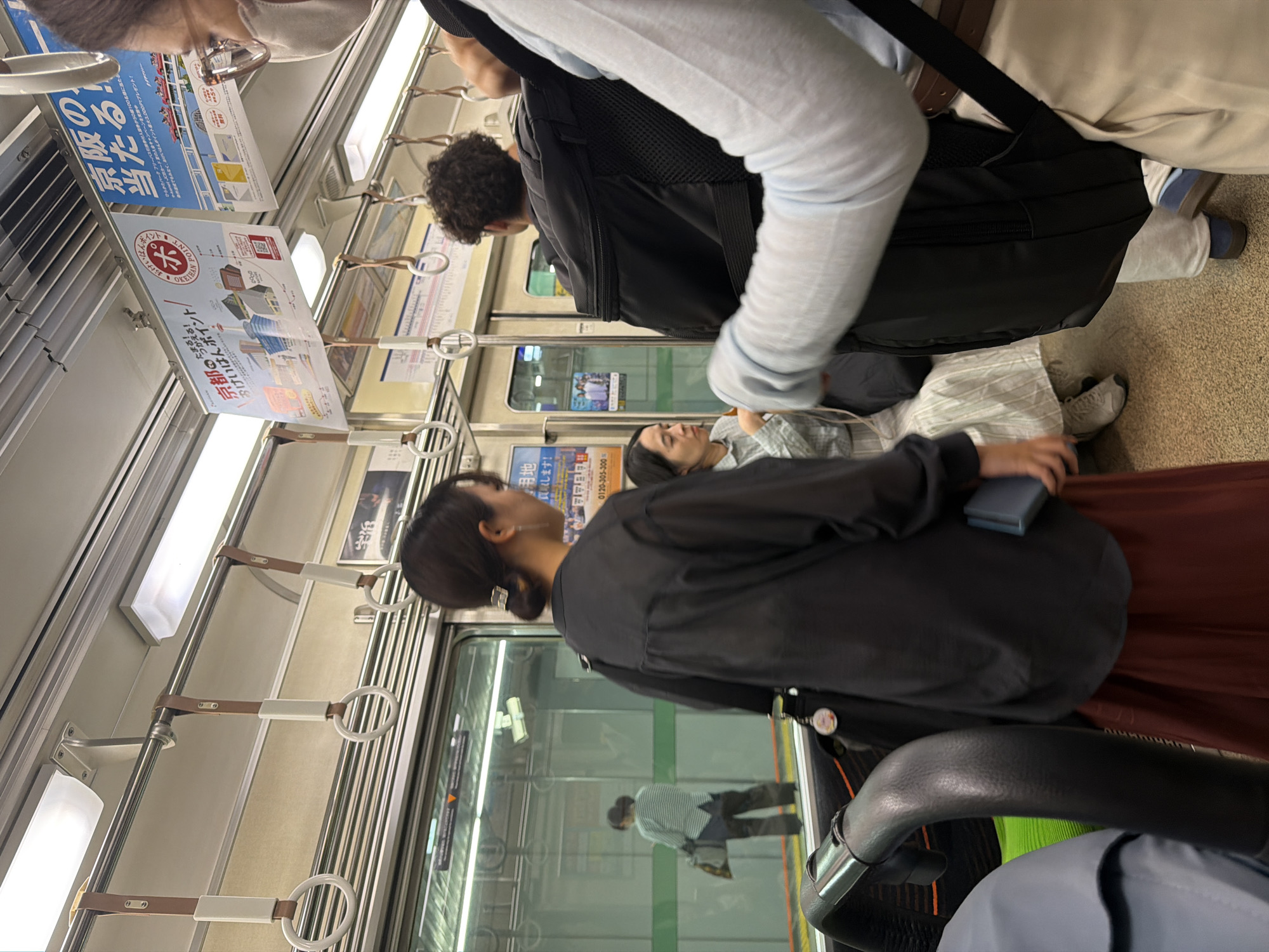The image depicts the interior of a passenger train car on the Keihan Line in Japan, specifically within the Kansai region, indicated by Japanese advertising posters for "Keihan" and "Kyoto" points displayed overhead.
In the immediate foreground on the right, the upper back and right side of a person are partially visible. This individual wears a light-colored long-sleeved top, light-colored lower garments, eyeglasses, and a light-colored face mask, carrying a dark backpack. A black, padded handrail curves into the lower-left foreground.
Multiple passengers are present in the mid-ground. On the left, a woman with dark hair tied in a low ponytail faces away from the camera, looking towards the left. She wears a dark, loose-fitting long-sleeved shirt and a long reddish-brown skirt, holding a small blue object in her left hand and carrying a dark shoulder bag with a circular charm. To her right, a person with dark, curly hair is seen from behind, wearing a dark shirt. Further right, a woman is seated on a bench, facing right and looking down at her hands. She has dark hair and wears a light-colored, possibly striped, long-sleeved shirt.
The train's interior features a series of overhead horizontal handrails with white rings and brown straps. Rectangular fluorescent light fixtures are mounted on the ceiling, illuminating the car. Various Japanese advertising posters, including one detailing "Keihan Point" with illustrations of Kyoto landmarks and another promoting "Keihan," are affixed to the ceiling panels. The floor has a textured, light-colored surface.
Through the train's window on the left, a reflection reveals a woman standing on an adjacent station platform, dressed in a light-colored horizontal striped shirt and dark pants, holding a light-colored bag. Green horizontal safety markings on the platform edge are also discernible in the reflection. The train's automatic sliding doors are visible behind the standing passengers in the mid-ground.