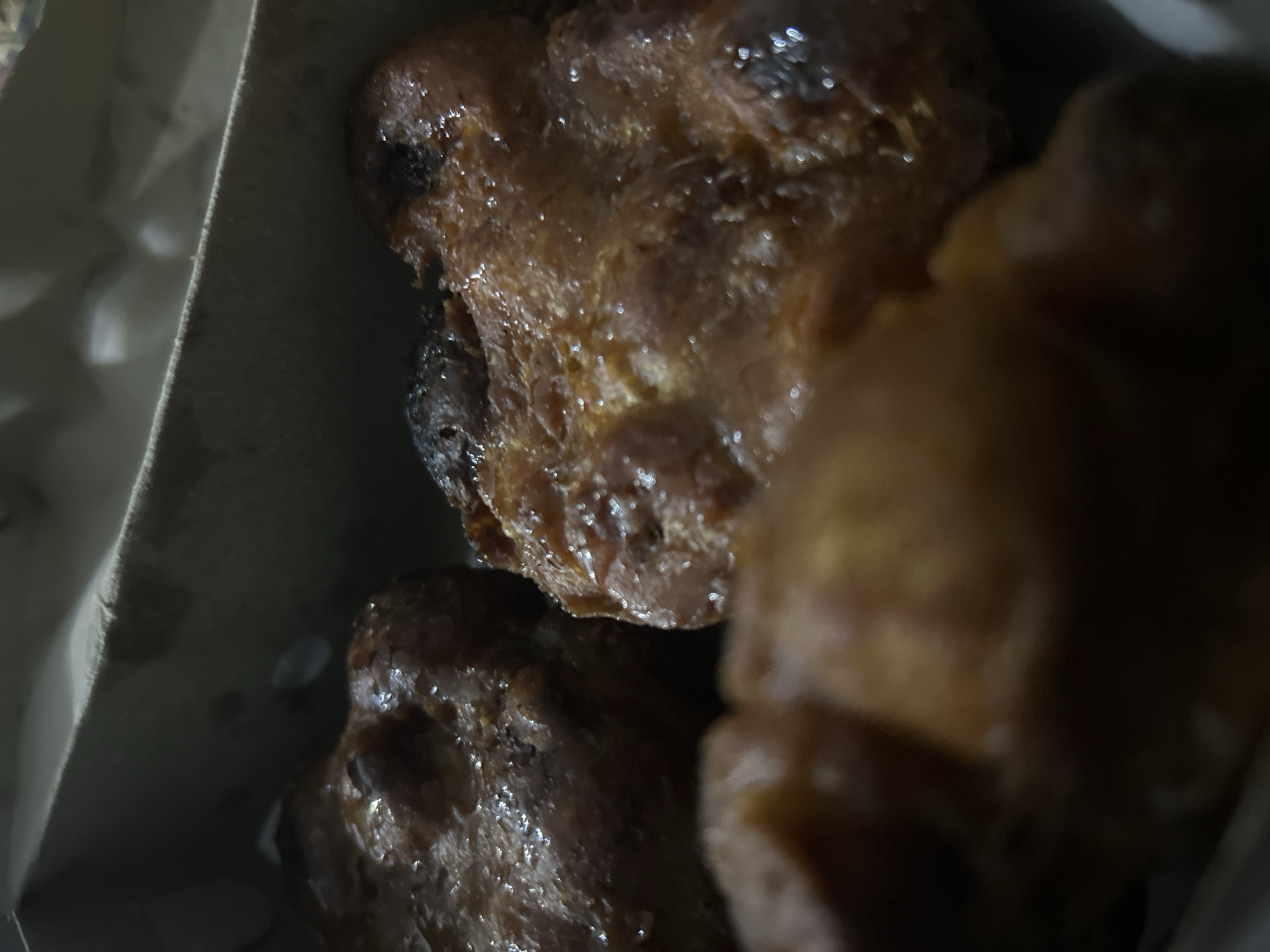 The image shows a close-up view of several fried dough pastries, likely "pisang goreng" or banana fritters, in a cardboard container. The pastries are golden brown with a slightly crispy texture, and some appear to be glazed or coated in sugar. The lighting is dim, suggesting the photo was taken indoors or at night. The background is dark and out of focus, with a hint of a light-colored paper or cardboard lining of the container. There is no visible text in the image. The setting appears to be a casual food vendor or a home kitchen in Sukawati, Indonesia. The overall impression is of a simple, appetizing snack.