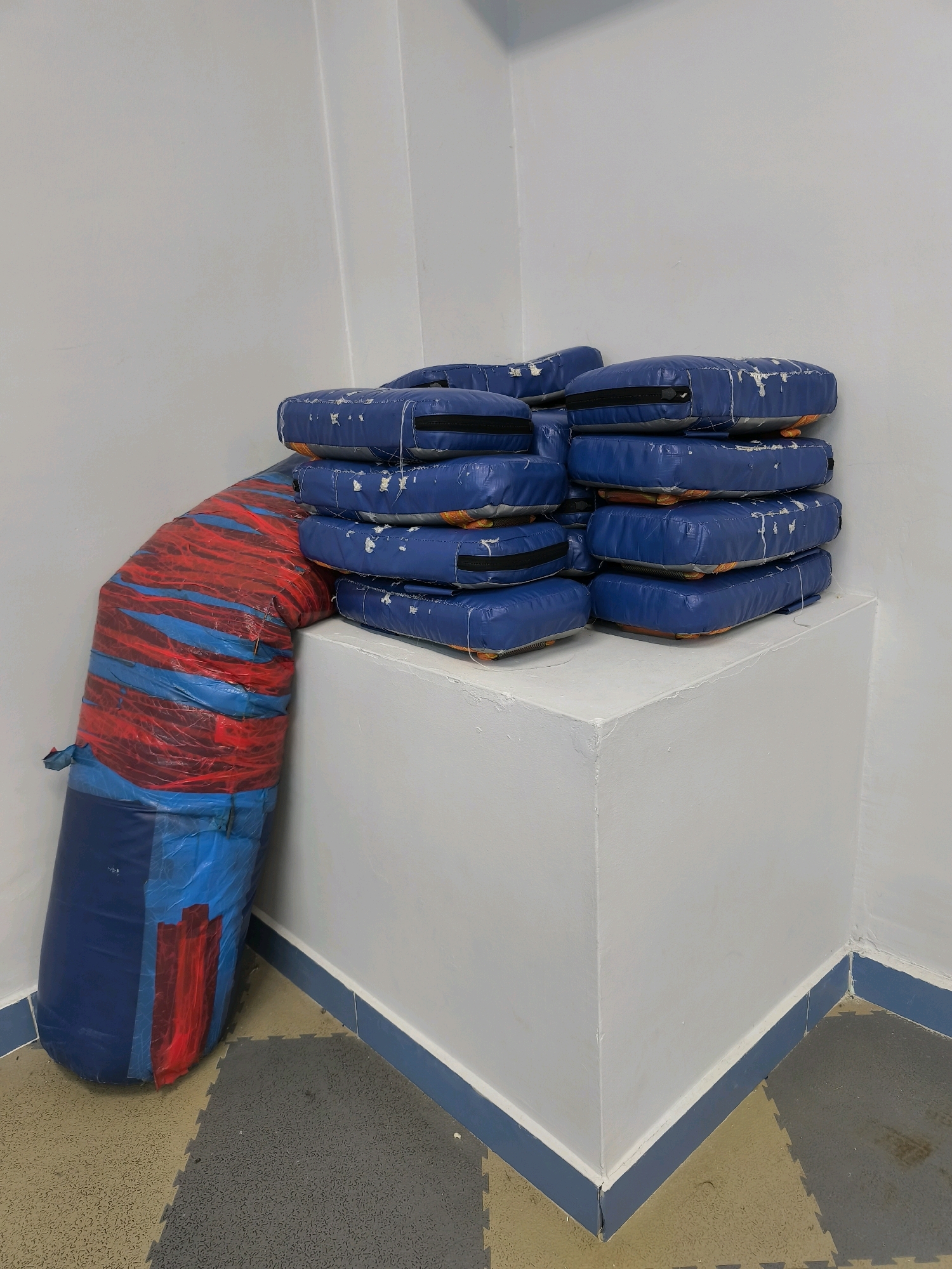 The image shows a corner of a room with two stacks of objects. One stack, on the right, is composed of rectangular blue objects, likely packages, stacked on top of each other. They appear to be wrapped in blue material with orange accents and have visible zippers. To the left of this stack, a large, cylindrical object is leaning against the wall. This object is covered in blue and red plastic wrap, creating a striped pattern. The room has white walls and a floor with interlocking grey and tan mats. The baseboard is painted blue. The lighting suggests an indoor environment, possibly a storage area or a warehouse. The image does not contain any information indicating its location in the city of Karachayevsk, Russia.