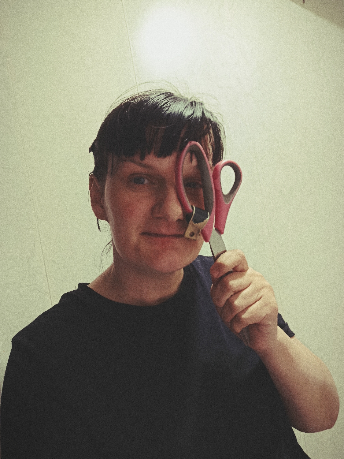 A close-up portrait features a person with short, dark hair and bangs, looking directly at the viewer. They are wearing a dark blue or black t-shirt. The person holds a pair of pink-handled scissors up to their face, with one of the finger holes positioned over their right eye (viewer's left). Their left eye is clearly visible, maintaining eye contact, and their mouth displays a subtle, neutral expression that might be a slight smirk. The metallic blades of the scissors are visible, and a section of one blade near the pivot appears to have some brownish or gold-colored tape or wear. The background is a plain, light-colored wall, possibly off-white or pale green, with a faint vertical line or texture. The lighting is soft and somewhat dim, with a slightly brighter area towards the top of the frame, giving the image a somewhat vintage or soft-focus aesthetic. There is no visible text. The scene is likely indoors, possibly in a home setting.