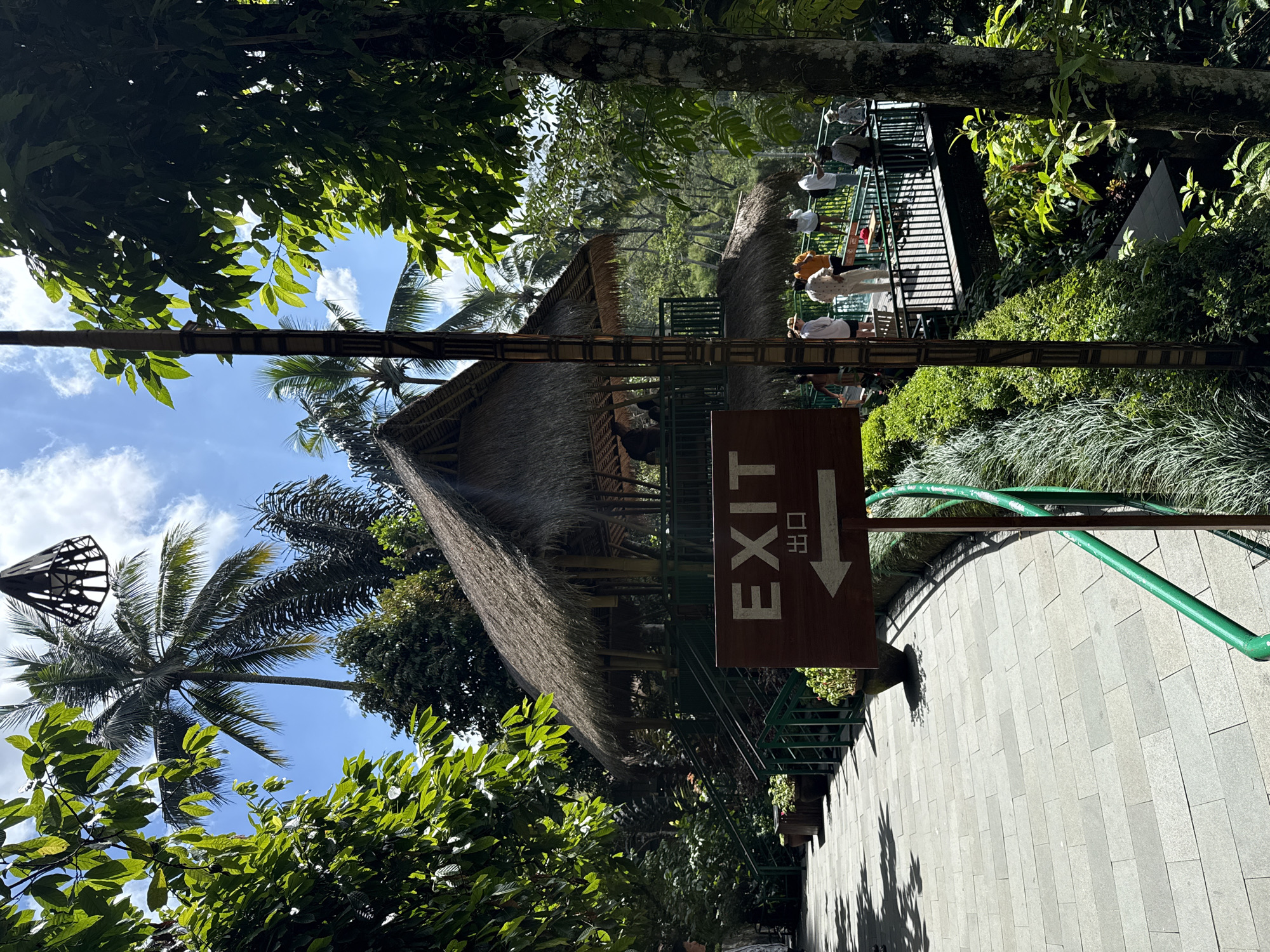The image depicts a tropical scene with lush greenery, palm trees, and a thatched-roof building, likely a rest stop or viewing area, set against a bright blue sky with scattered clouds. In the foreground, a wooden sign points left with the word "EXIT" and Japanese characters below it. A paved walkway curves to the left, bordered by a green railing. Several people are visible on a raised wooden platform in the background, some observing the surroundings. The environment suggests a tourist destination, possibly part of a jungle resort or park in Tegallalang, Indonesia, known for its rice terraces. The overall impression is one of a serene, natural setting enhanced by man-made structures for visitor comfort and guidance.