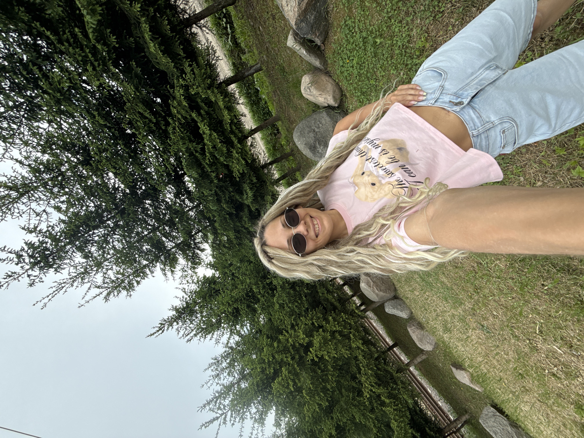 A young woman with long blonde hair is lying on a grassy lawn. She is wearing sunglasses, a pink t-shirt with a teddy bear graphic, and light blue jeans. The lawn is surrounded by trees and stones, which form a makeshift border. The scene appears to be in a park or green space, with a cloudy sky visible in the background. The location may be Rusanovo, Russia.