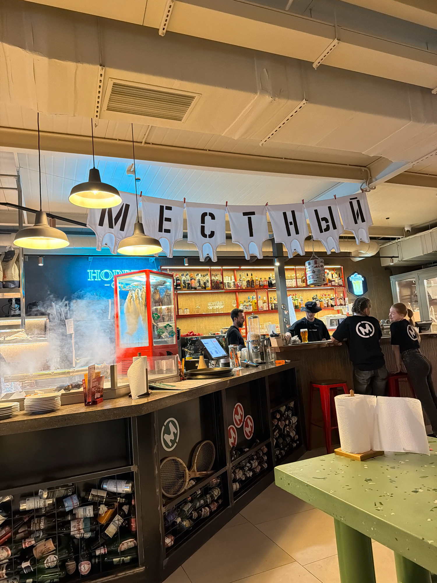 The image depicts the interior of a restaurant or bar, likely located in Gelendzhik, Russia, as suggested by the banner featuring Cyrillic lettering. The banner, composed of white t-shirts with black letters, reads "МЕСТНЫЙ" which translates to "LOCAL". Several people are visible behind a counter, appearing to be staff. One person is wearing a black hat and serving drinks, while others are in black t-shirts with a circular logo. Two individuals are conversing at the counter. The environment is illuminated by pendant lights and neon signage that reads "HOHDE". A refrigerated display case with hanging cured meats is prominently featured. The counter area is stocked with bottles of alcohol and glassware, and the shelves behind the bar are also filled with various bottles. In the foreground, a green table with a roll of paper towels suggests a dining area. The overall impression is a casual establishment with a focus on local products or atmosphere.