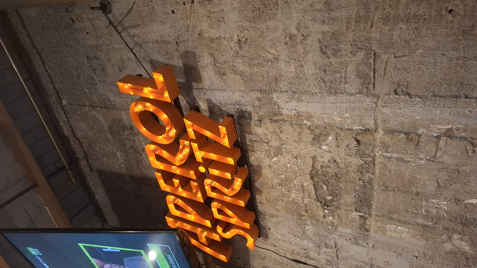 This upward-angled photograph, captured in Sofia, Bulgaria, depicts a section of an interior wall displaying an illuminated sign and a television screen. The central subject is a three-dimensional, orange-colored sign spelling "APEROL SPRITZ" in two lines, with "APEROL" positioned above "SPRITZ." The letters are internally lit by visible, warm-toned light bulbs, casting subtle shadows on the wall behind them. The sign is mounted on black brackets, and a black electrical cable is visible extending to the left from the "APEROL" section.
The wall exhibits a rough, textured surface, appearing to be constructed from dark grey and brown concrete or stone blocks with varying textures and patches of lighter, possibly peeling, material. To the upper left of the image, a television screen is mounted, displaying a live broadcast. Visible text on the screen includes "SPORT," "HD," and "LIVE," along with a partial view of a person and some Cyrillic text at the bottom, reading "БЪЛГАРИЯ АРМИЯ" (Bulgaria Army). Above the sign and to the right, a thin, metallic pipe or conduit runs horizontally across the upper portion of the wall. Further to the right of the sign, a dark metal hook or bracket is affixed to the wall. The overall environment suggests an establishment with an industrial or rustic aesthetic.