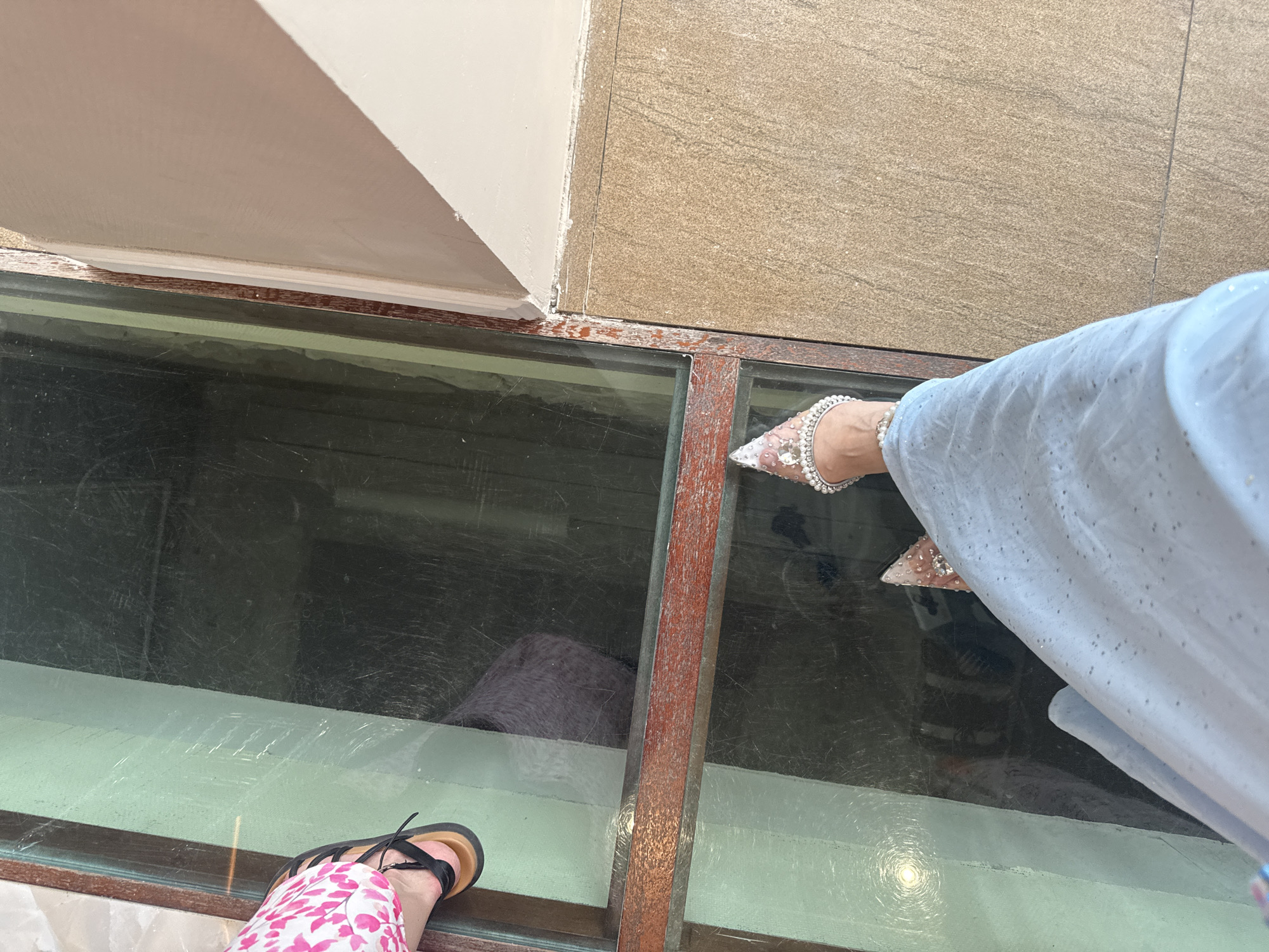The image captures a low-angle, personal perspective of two individuals standing on a transparent floor, likely glass, which looks down onto another level. The person in the foreground is wearing a light blue, shimmering dress and an ornate, bejeweled shoe. Their left leg is visible, extending down and to the left. The second person, on the left side of the frame, is wearing a pink and white patterned garment and black sandals. Their feet are positioned over the glass floor, with one foot partially visible. The flooring beneath the glass appears to be a dark, possibly reflective surface. The glass floor is framed by what seems to be a weathered wooden structure. Adjacent to the glass floor is a textured, light brown tiled surface. The overall environment suggests an indoor space with a unique architectural feature. The image does not contain any visual cues or context that would allow for the determination of its location in Komsomolsk-on-Amur, Russia.