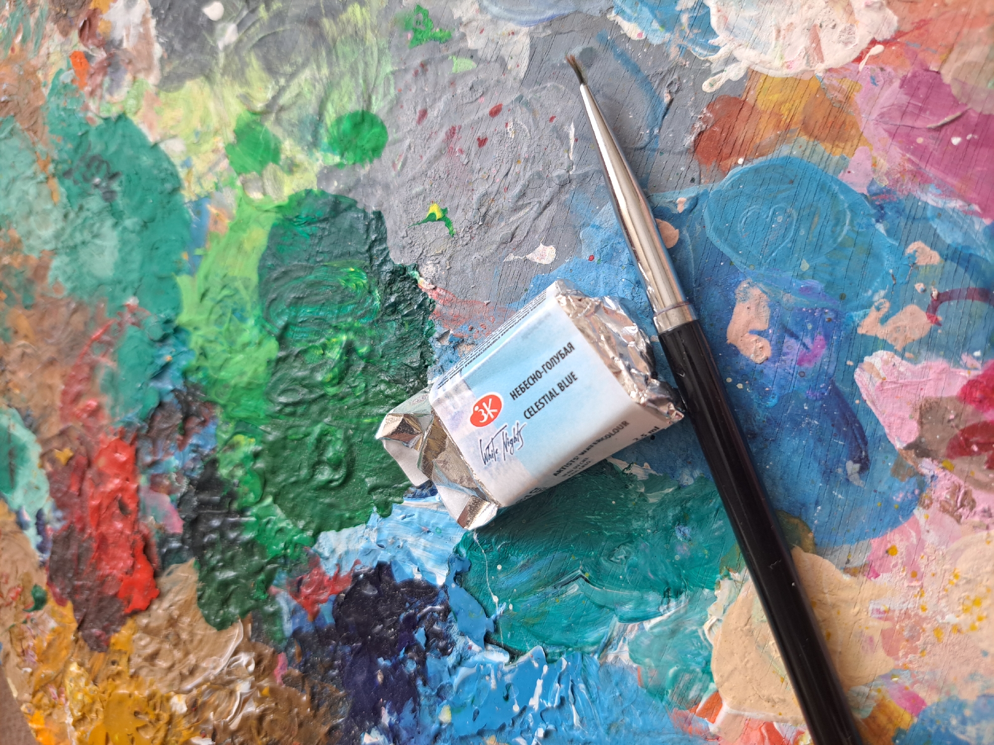 This is a close-up, overhead shot of an artist's palette laden with thick, colorful paint. The palette itself appears to be made of wood and shows signs of extensive use, with dried paint layered upon dried paint in a vibrant mosaic of blues, greens, yellows, reds, and purples. A silver-tipped paintbrush with a black handle rests diagonally across the palette, its bristles slightly splayed.
In the center of the image, a small, foil-wrapped watercolor paint pan is open, revealing a light blue pigment. The label on the paint pan is partially visible and reads "3K" within a red circle logo, followed by Russian text "НЕБЕСНО-ГОЛУБАЯ" and "CELESTIAL BLUE" in English. Below this, it indicates "ARTISTS WATERCOLOUR" and "3.5 ml". The paint pan is positioned as if recently used or about to be used.
The overall impression is one of a working art studio, possibly in Popovo, Bulgaria, where an artist is in the process of creating. The textures of the paint are prominent, suggesting an impasto technique or simply the build-up of layers over time. There are no people visible, but the presence of the tools and materials strongly implies an artistic activity. The lighting is bright and natural, suggesting daytime. The emotional tone is one of creativity and artistic exploration.