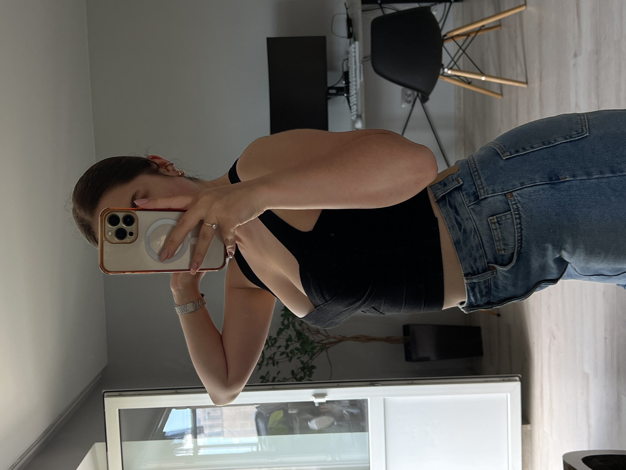 A woman is standing in a room, holding a smartphone to take a selfie in a mirror. She is wearing a black, sleeveless crop top and light-wash denim jeans. The room appears to be a home office or living space, with a desk, computer monitor, keyboard, and a black chair visible in the background. A large, white-framed window or door is to the left of the frame, and a potted plant is situated between the woman and the window. The floor is light-colored wood. The image does not contain any information that would allow for its location to be identified as Ternopil, Ukraine.