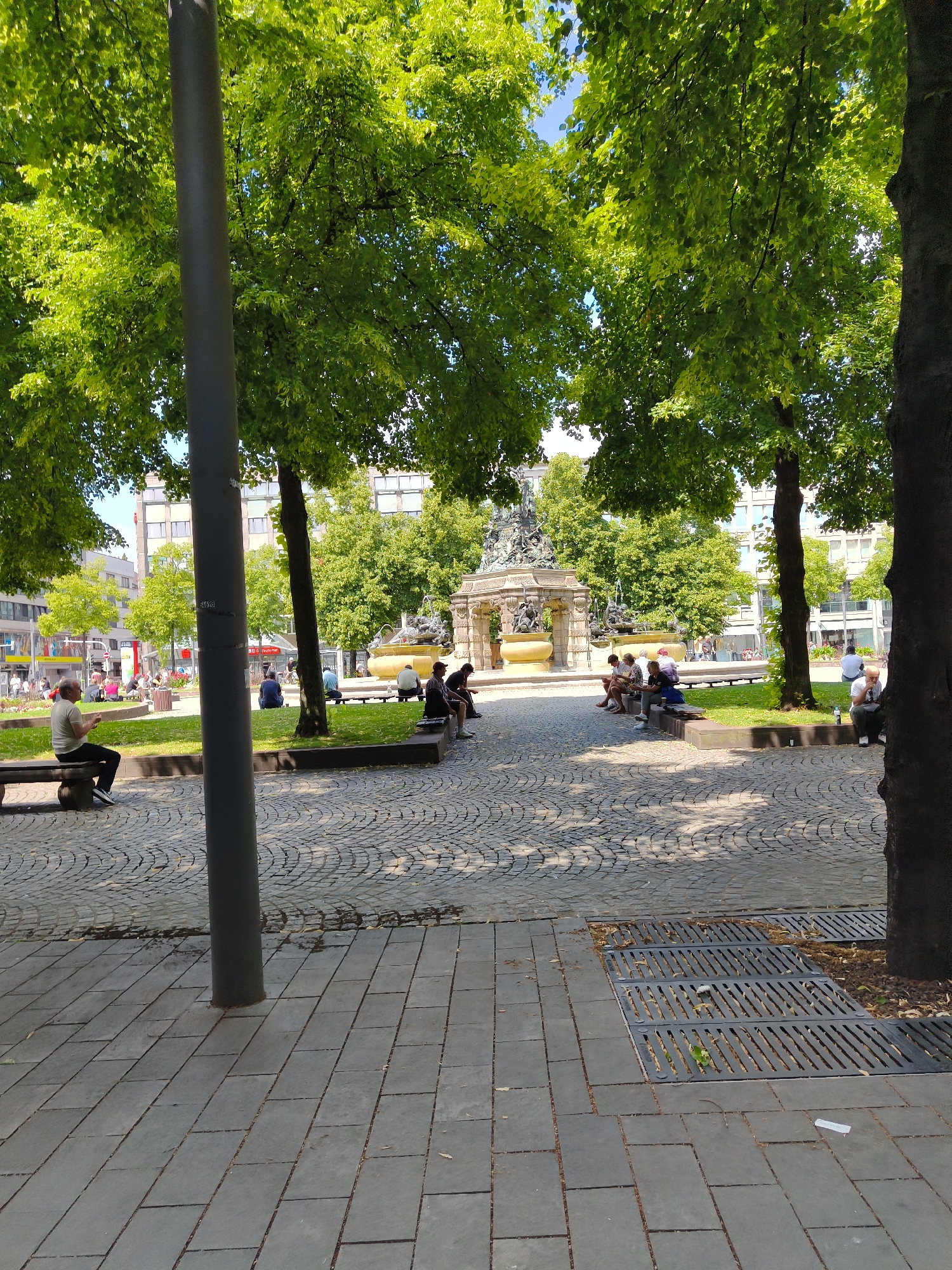 The image depicts a public square in Mannheim, Germany, likely Marktplatz, given the presence of the Hercules Fountain. The scene is bathed in sunlight, casting dappled shadows from the mature trees that line the square. Several people are seated on benches and along the low walls surrounding a grassy area.
In the foreground, a dark gray lamppost stands prominently. The ground is paved with interlocking gray stones, transitioning to a coarser, circular pattern of cobblestones. Several metal grates, likely for drainage, are visible in the paved area.
To the left, a man in a light-colored shirt and dark pants sits alone on a wooden bench, looking at something in his hands. Further back, a group of people are gathered on benches in the grassy area.
The centerpiece of the square is the Hercules Fountain, a large, ornate monument with sculptures and water features. The fountain is surrounded by a low, yellowish-brown stone wall.
In the background, modern buildings with multiple windows and balconies form the backdrop to the square. Some shops and street signs are visible on the ground floors of these buildings. The overall atmosphere is one of a relaxed, sunny day in an urban public space.