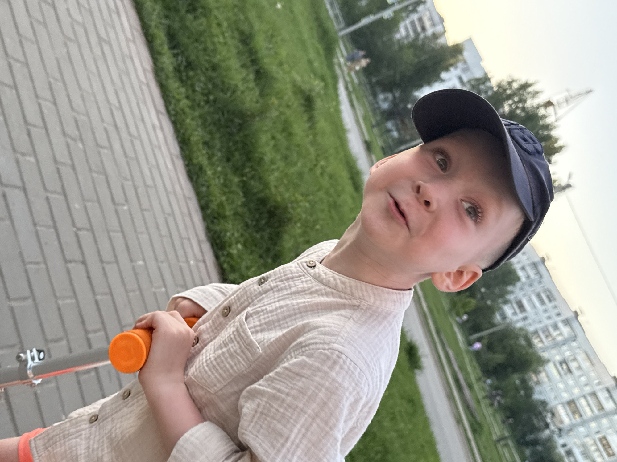 A young boy stands on a paved area, possibly a sidewalk, with greenery and apartment buildings in the background. He is wearing a light-colored, button-up shirt and a dark blue baseball cap. He holds an orange scooter handle. The location may be in the city of Chaikovsky, Russia. The angle of the shot is canted, making the image slightly tilted. The setting appears to be outdoors during daylight hours.