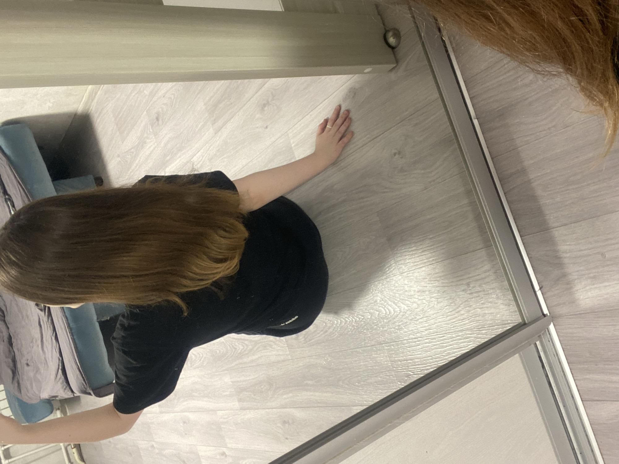 This image captures an indoor scene, likely within a residence in Chaikovsky, Russia, taken from a high vantage point, possibly a mirror selfie. The main subject is a person, likely a woman with long, wavy brown hair, wearing a black short-sleeved top and black bottom wear. Their right arm is extended, and their hand is resting on a light-colored, wood-grain patterned floor. A thin, possibly gold, band ring is visible on the fourth finger of their right hand.
The floor covers most of the visible area, exhibiting a consistent light greyish-white wood laminate or vinyl pattern. In the background, to the left of the person, a piece of light blue upholstered furniture is visible, partially obscured by a grey textile or blanket. Above the blue furniture, a light-colored wooden or laminate structure, possibly a shelf, bed frame, or part of a closet, runs horizontally. To the right, a silver metal track, likely for a sliding door or mirror, is prominent.
The photo itself appears to be a reflection, as indicated by the photographer's own hair visible in the upper right corner of the frame. On the person's black clothing, faint white text, partially visible, reads "adibo", suggesting the "Adidas" brand logo. The overall setting appears modern and uncluttered. No specific time of day or emotional state can be determined from the image.