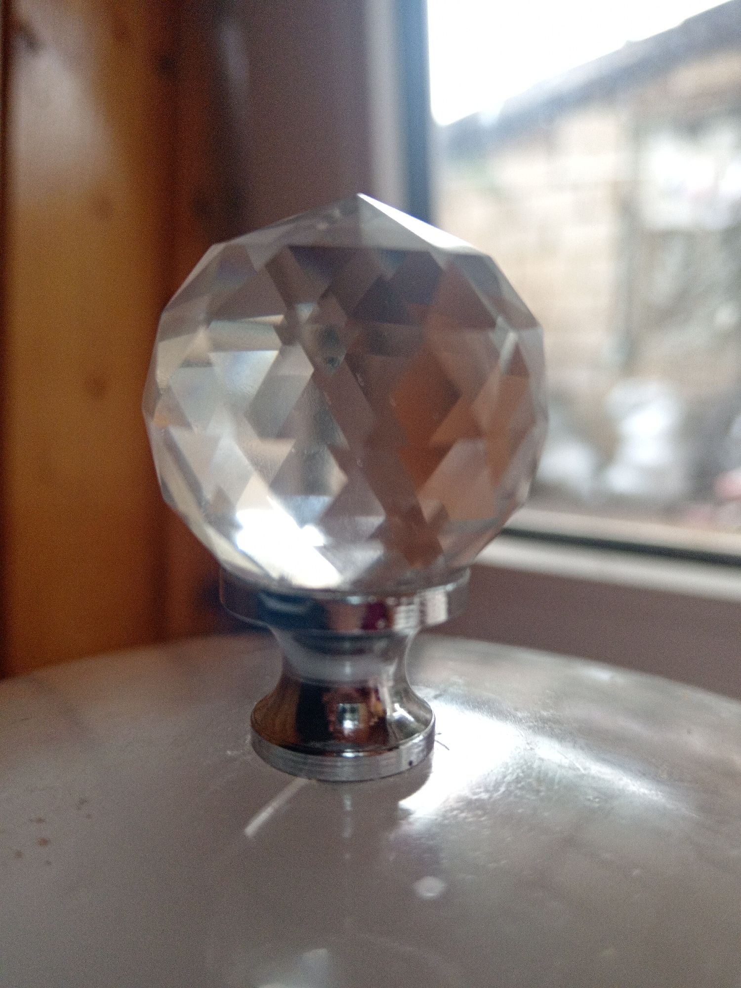 A faceted crystal knob, likely attached to a lid or drawer, is the central focus of the image. The knob is clear, refracting light and reflecting its surroundings. It is mounted on a metallic base, which appears to be silver or chrome, with a decorative, flared shape. The knob sits atop a light-colored, smooth surface, possibly ceramic or enameled metal. In the background, a wooden surface is visible on the left, and a blurred window on the right looks out onto an outdoor scene with indistinct structures and foliage. The overall lighting suggests an indoor setting, possibly near a window. The image does not contain any people. The location within the city of Uchkeken, Russia, cannot be determined from the visual information provided.