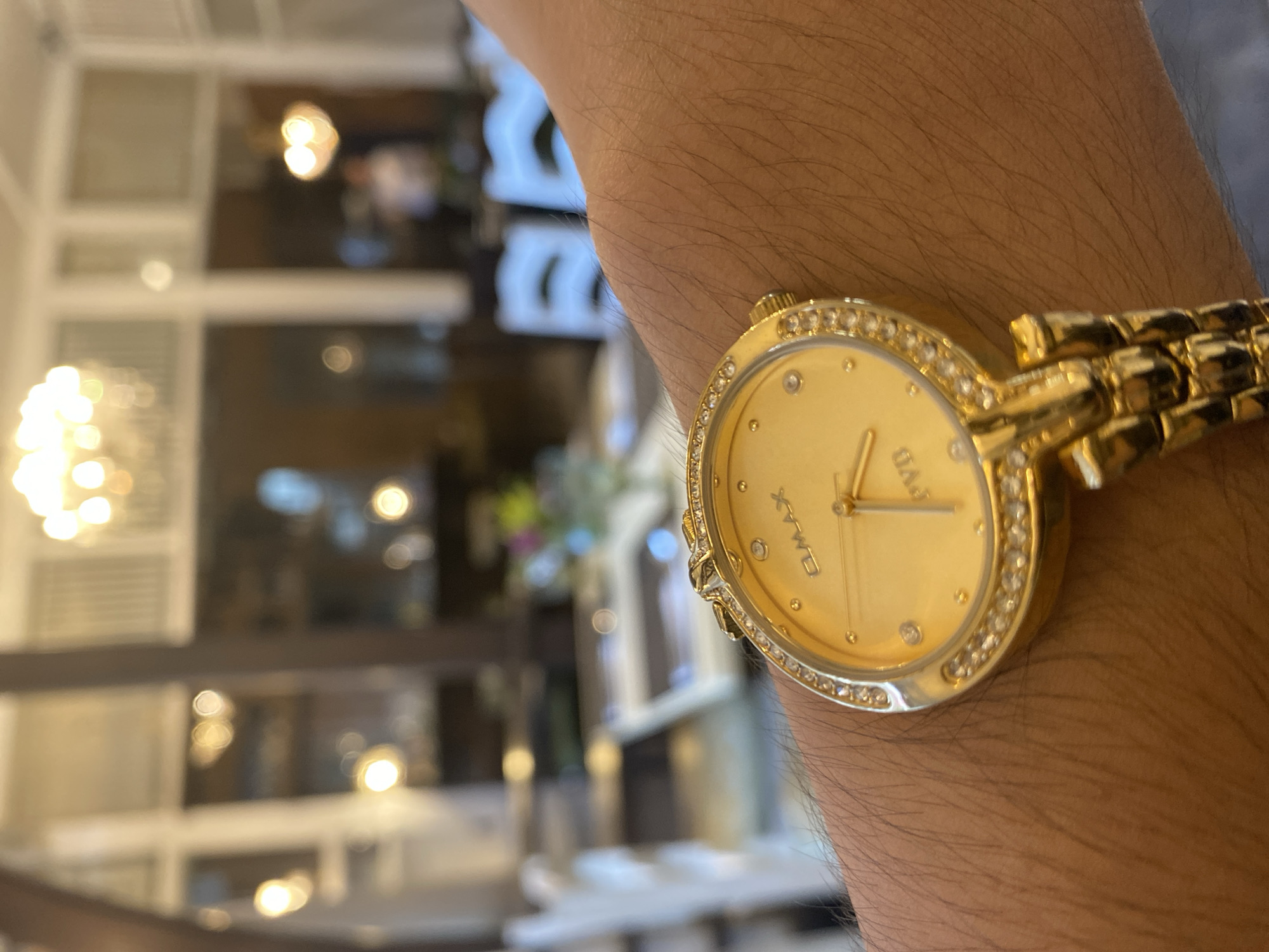 The image is a close-up of a gold OMAX watch with diamond accents on a person's wrist. The watch face is gold with a second hand. The background is out of focus and appears to be an indoor dining setting with tables and chairs, illuminated by chandeliers. The environment suggests a restaurant or a formal dining area. There are no visible identifying landmarks or clues to definitively place the location in Davao City, Philippines.