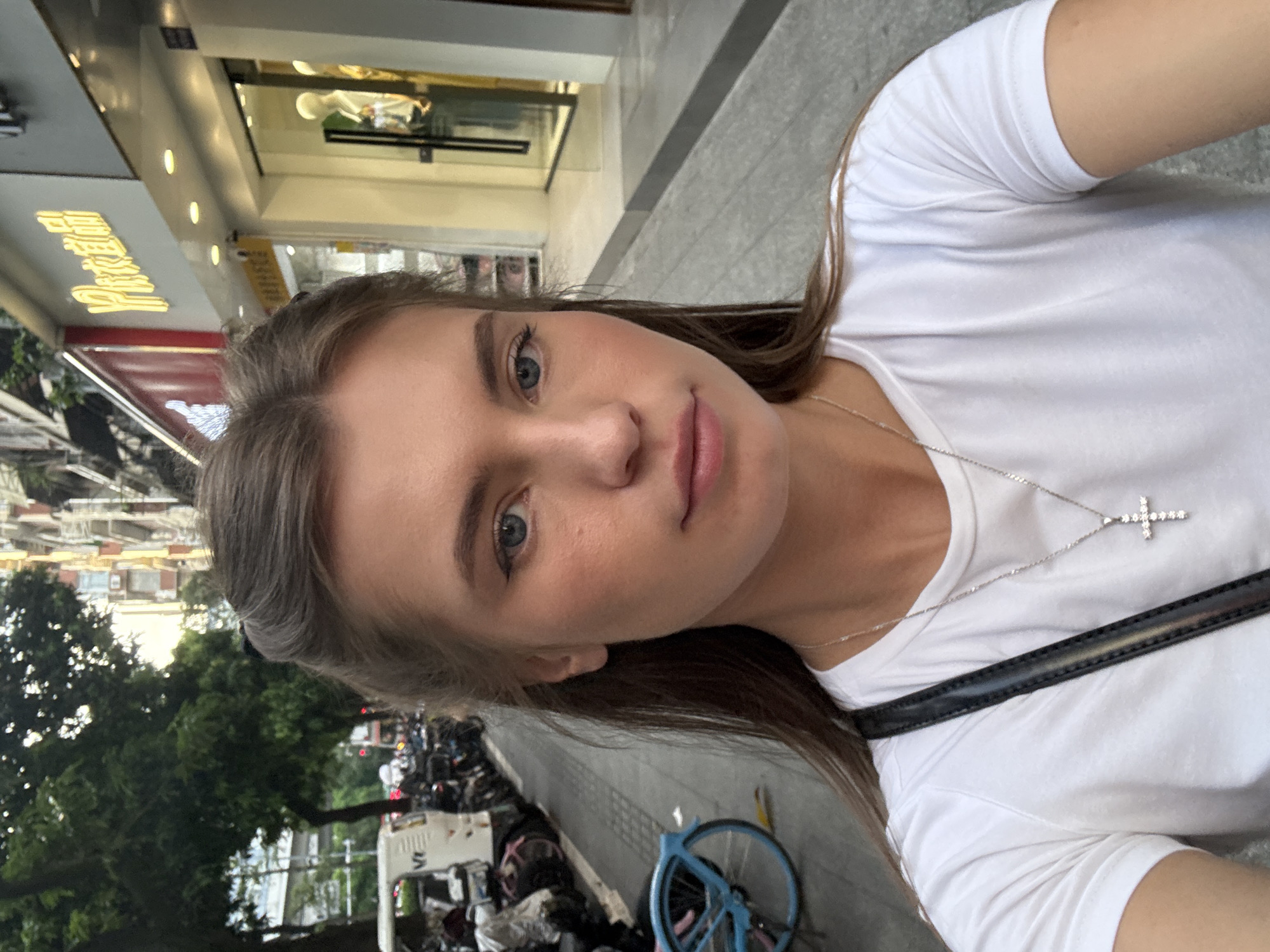 The image is a medium shot selfie of a young woman standing on a sidewalk in Haizhu District, China. She is wearing a white t-shirt and a silver cross necklace. Her hair is pulled back on the sides, with the rest falling loose. She is looking directly at the camera with blue eyes and light makeup.
Behind her, the sidewalk is lined with parked motorcycles and a blue bicycle. A white van is visible to the left, and shops with illuminated signs are on the right. The buildings appear to be multi-story structures with shops on the ground floor. The lighting suggests it is daytime, possibly late afternoon, with a mixture of natural light and artificial illumination from the shops. The overall scene depicts a typical urban street in a commercial area.