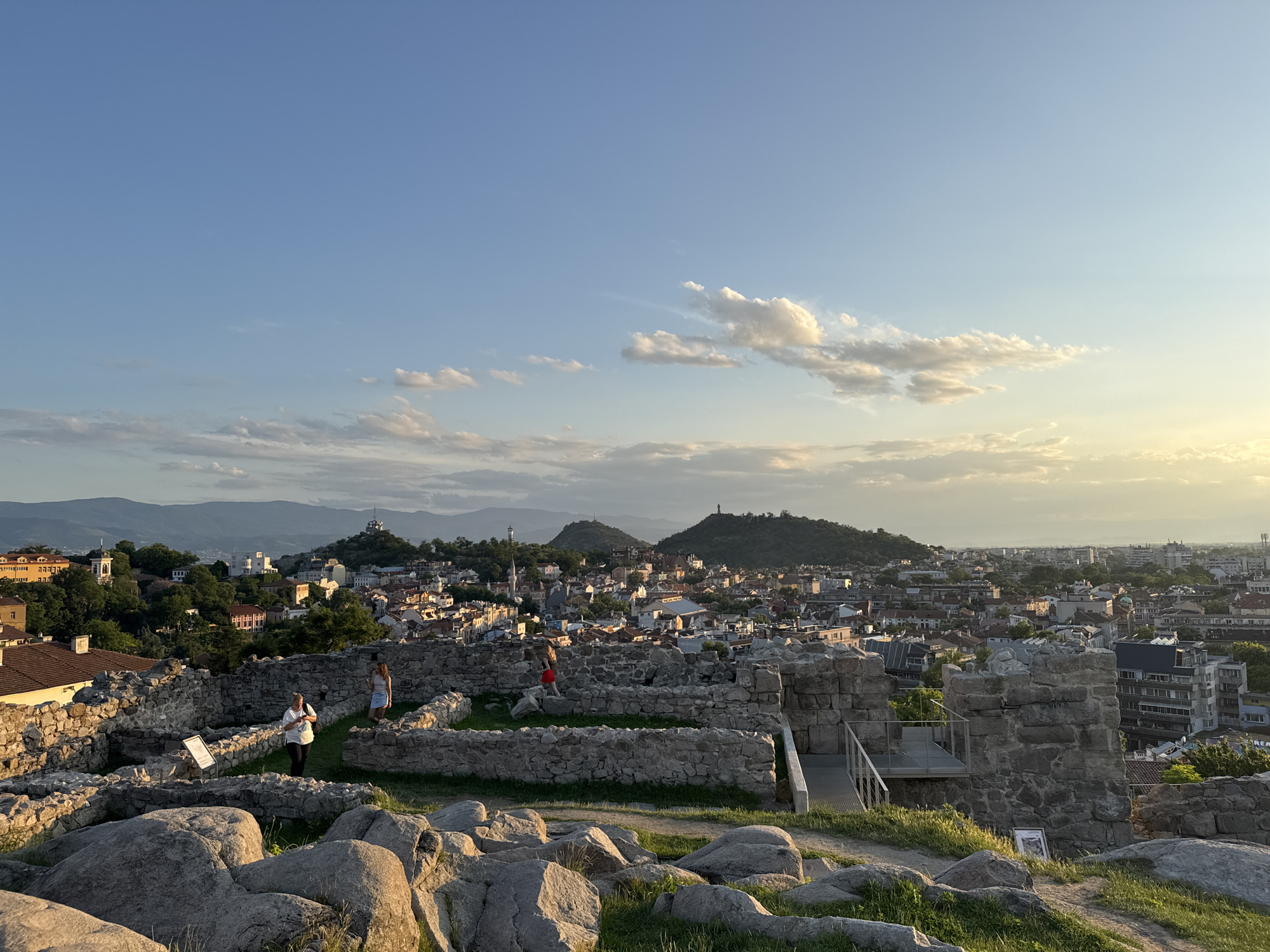 The image presents an elevated panoramic view of the city of Plovdiv, Bulgaria, from an archaeological site, likely Nebet Tepe hill, during the late afternoon or early evening. Golden sunlight enters the frame from the right, illuminating the scene.
In the foreground, large, irregularly shaped grey boulders and patches of green grass cover the terrain. Beyond these, a network of grey stone walls, representing ancient ruins, forms distinct enclosures and pathways. Three individuals are present within these ruins: one woman, dressed in a white shirt and dark trousers, stands on the left side of the ruins; a second woman, wearing a light-colored top and skirt, stands slightly behind and to the right of the first woman; a third person, in a red top and dark trousers, walks along a grassy path within the ruins further to the right. A modern metal walkway or viewing platform is visible on the right side of the ruin complex.
The midground showcases the dense urban landscape of Plovdiv. Numerous buildings, many with terracotta-tiled roofs, are closely packed and interspersed with green trees. Several hills rise within the cityscape. In the center-right, two prominent hills are visible, each topped with a distinct structure. The left of these two hills features a spire-like edifice, while the rightmost hill is crowned by a large commemorative statue, identifiable as the Alyosha Monument on Bunardzhik Hill.
The background consists of a distant, undulating range of blue-hued mountains stretching across the horizon. The sky overhead is a clear light blue, transitioning to a bright golden-yellow on the right horizon, where scattered white and grey cumulus clouds are visible, illuminated by the low-angle sunlight.