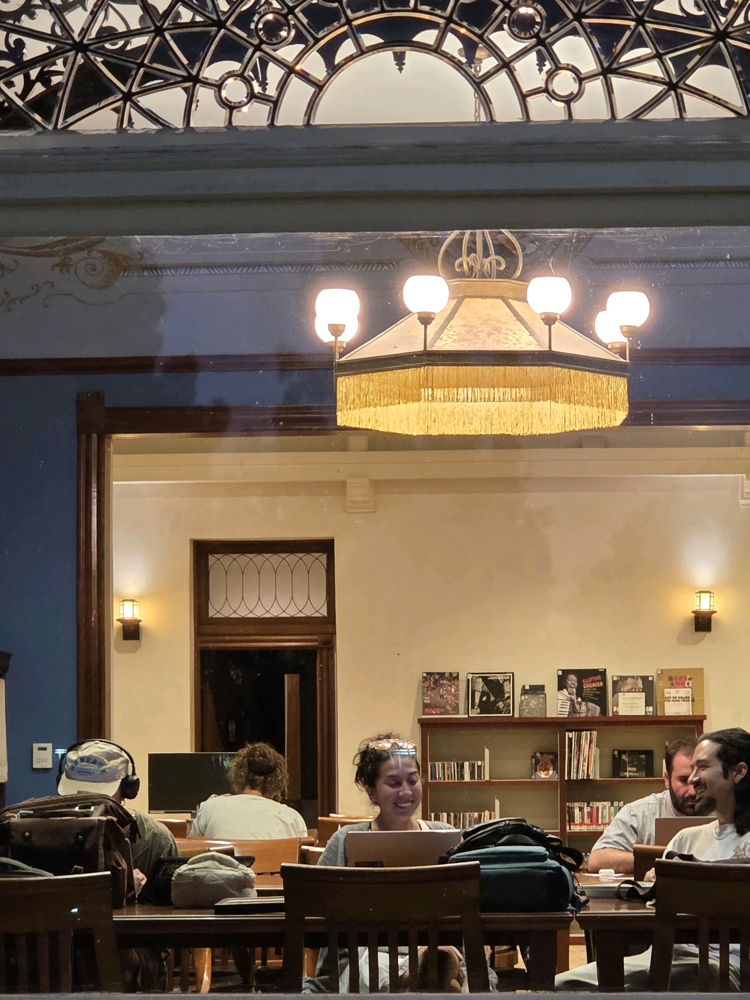 The image depicts an indoor scene, likely a library or a study hall, within the city of Glendale, United States of America. The setting features wooden furniture, bookshelves filled with books and records, and ornate architectural elements including a stained-glass ceiling and a large, tasseled chandelier. Several individuals are present, engaged in studying or working on their laptops. One person in the foreground wears headphones and a cap, while another, a woman, is smiling and looking at her laptop screen, with what appears to be a digital overlay on her face. Two other individuals are visible in the background, also seemingly focused on their work. The ambiance suggests a quiet, studious environment.