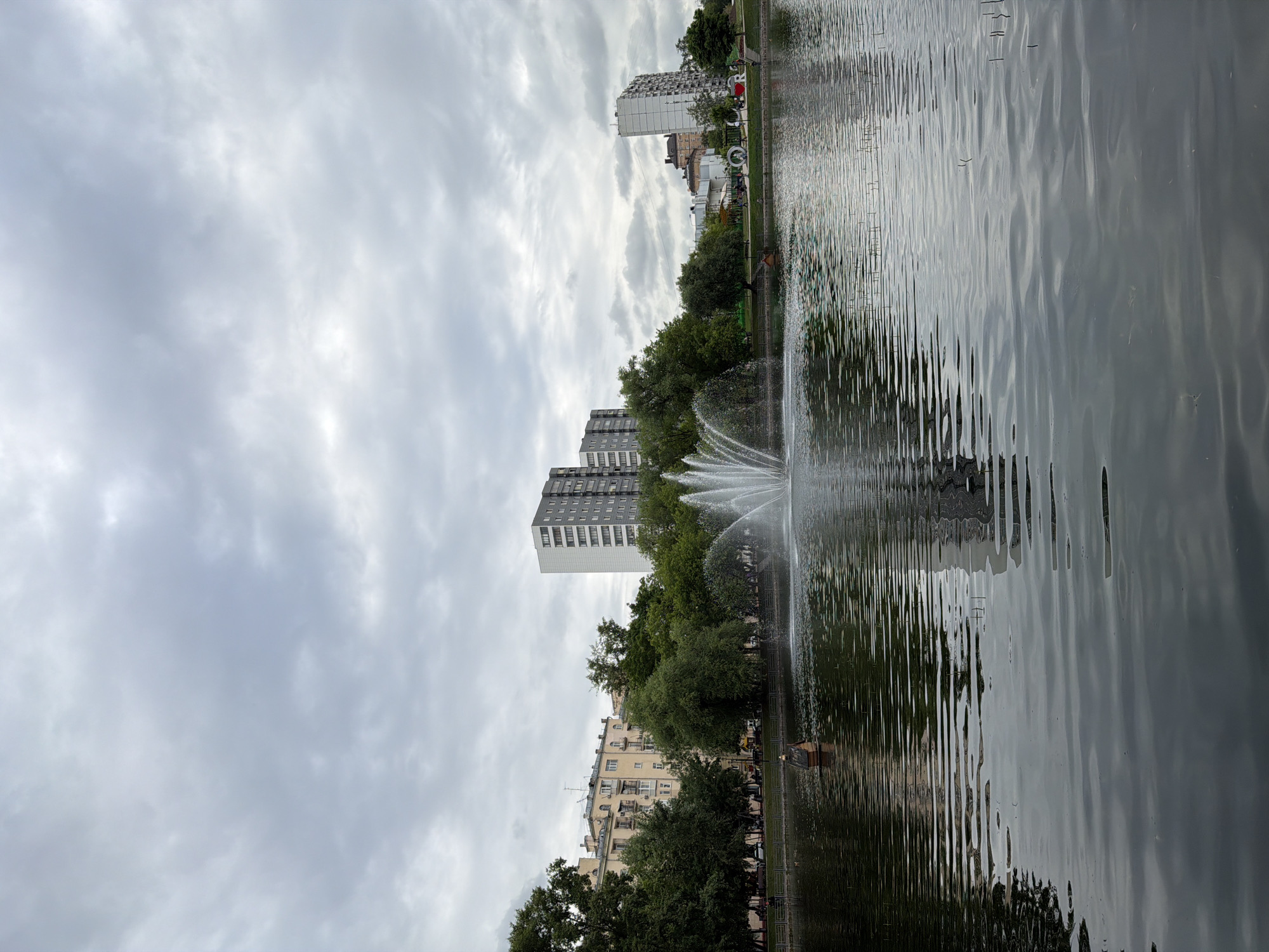 A serene view of a park with a lake, fountain, and buildings under a cloudy sky. The lake, in the foreground, has a gentle ripple pattern on its surface, reflecting the overcast sky and the surrounding greenery. A fountain shoots plumes of water into the air in the center of the lake. On the bank of the lake, trees with lush green foliage line the perimeter, interspersed with several buildings. Two multi-story apartment buildings, one light-colored with many windows and balconies, and another taller, gray building with a grid-like facade, stand out against the sky. In the distance, other buildings of varying heights and architectural styles are visible. A small, wooden structure with a pitched roof sits on a small island in the lake. The overall atmosphere is peaceful and tranquil, characteristic of an urban park setting.