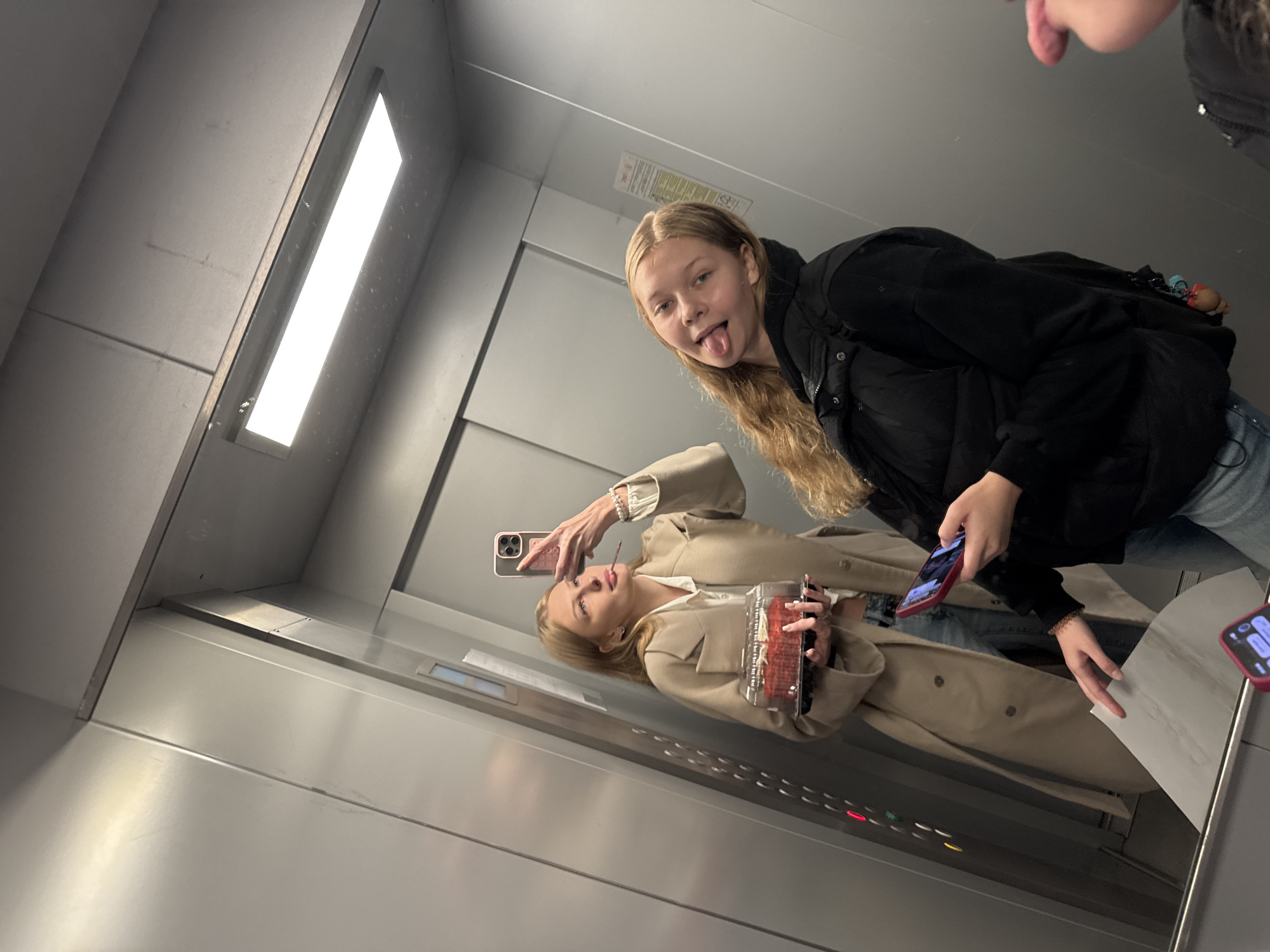 Two young women are posing for a selfie in an elevator, likely in an apartment building or commercial establishment. The elevator's interior is characterized by brushed stainless steel walls and a bright fluorescent ceiling light.
The woman on the left, with blonde hair, is holding a clear plastic container filled with red velvet cupcakes. She is wearing a beige trench coat over a white shirt and blue jeans. She is holding a pink cell phone and looking at it.
The woman on the right, also with blonde hair, is sticking her tongue out and looking at the camera. She is wearing a black puffer vest over a dark top and blue jeans. She is holding a red-cased cell phone.
The elevator control panel is visible on the left wall, featuring illuminated buttons. The presence of a bright, modern elevator suggests an urban setting. While the specific location within the city of Perm, Russia cannot be definitively identified from the visual cues alone, the image depicts a common urban environment.