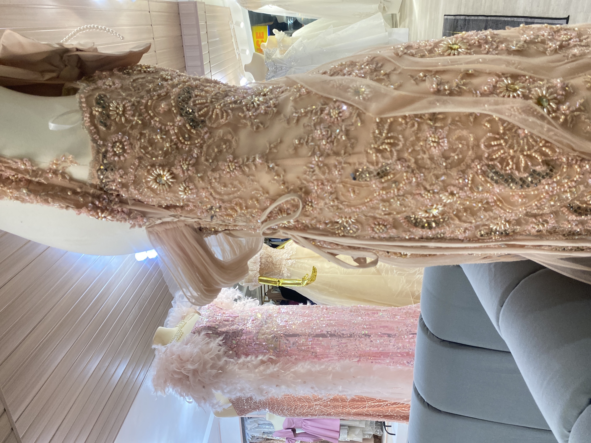 This image shows the interior of a formal wear boutique, likely a bridal or evening gown shop, located in Davao City, Philippines. The focal point is a mannequin displaying an intricately beaded and embellished gown in a peachy-pink hue. The gown features delicate floral and swirling patterns made of beads, sequins, and possibly pearls, with sheer sleeves and a trailing skirt.
Behind the main mannequin, other dresses are visible on display, including a pink sequined gown with a feathery neckline and a creamy yellow gown. Further back, white wedding gowns can be seen. The setting appears to be well-lit, with fluorescent lighting or spotlights illuminating the merchandise. A light blue upholstered sofa is partially visible in the foreground, suggesting a waiting or viewing area for customers. The ceiling is paneled with a light-colored material. The overall impression is one of elegance and opulence, showcasing a selection of formal attire.