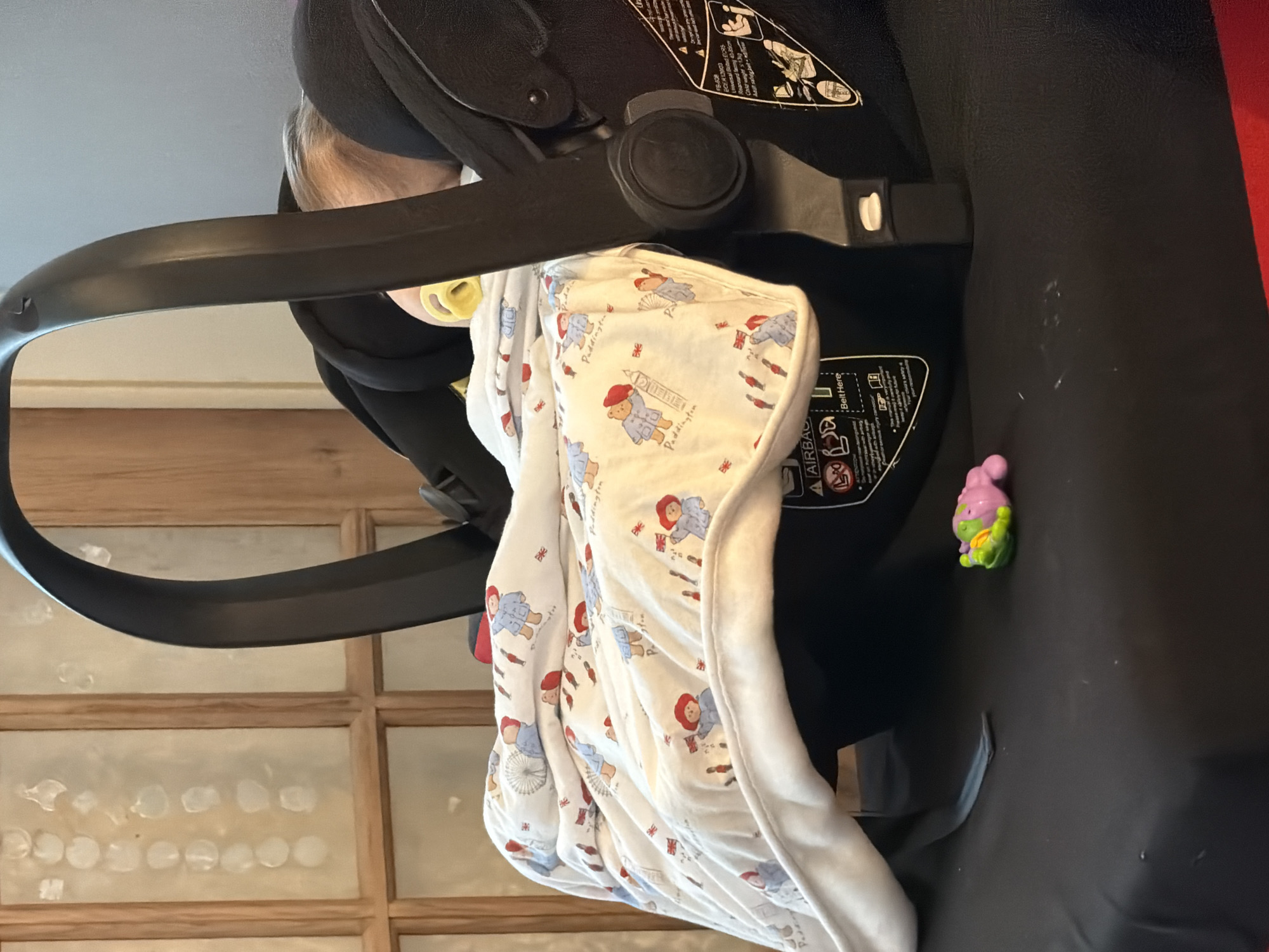 A baby, with a yellow pacifier in its mouth, is buckled into a car seat and covered with a Paddington Bear-themed blanket. A small toy is next to the car seat, against a black surface. The background shows a grid-patterned window. The car seat's information sticker is also visible.