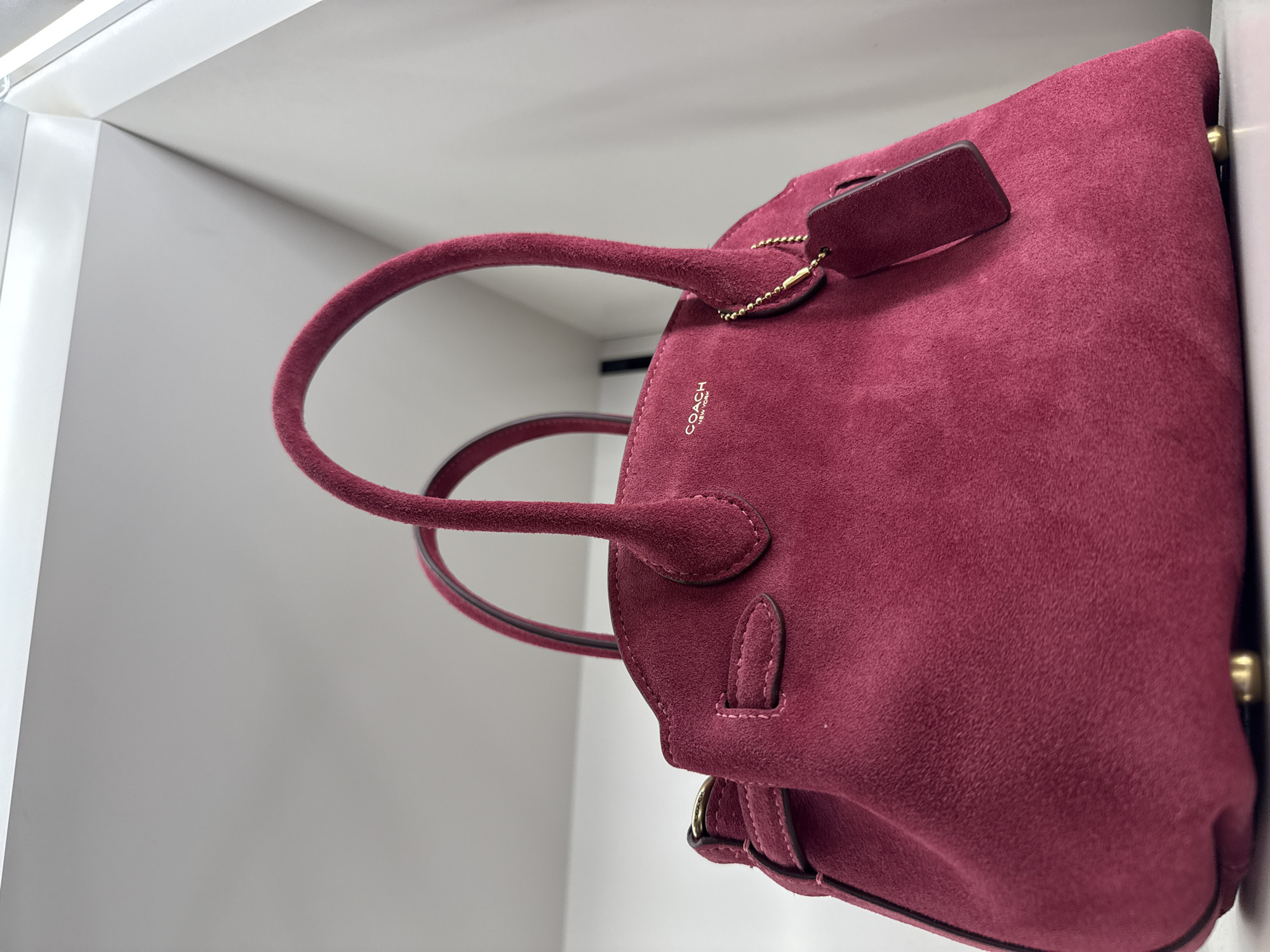 The image captures a close-up, low-angle shot of a maroon-colored suede handbag. The bag appears to be a Coach brand, with the logo subtly visible on the surface. The bag has two handles, and a small tag is attached. The bag is situated in a bright, presumably indoor, location, possibly a retail store, with white surfaces forming the background. The geographical location is identified as Sunrise, United States of America. No people or specific interactions are visible in the scene. The environment appears clean and well-lit.