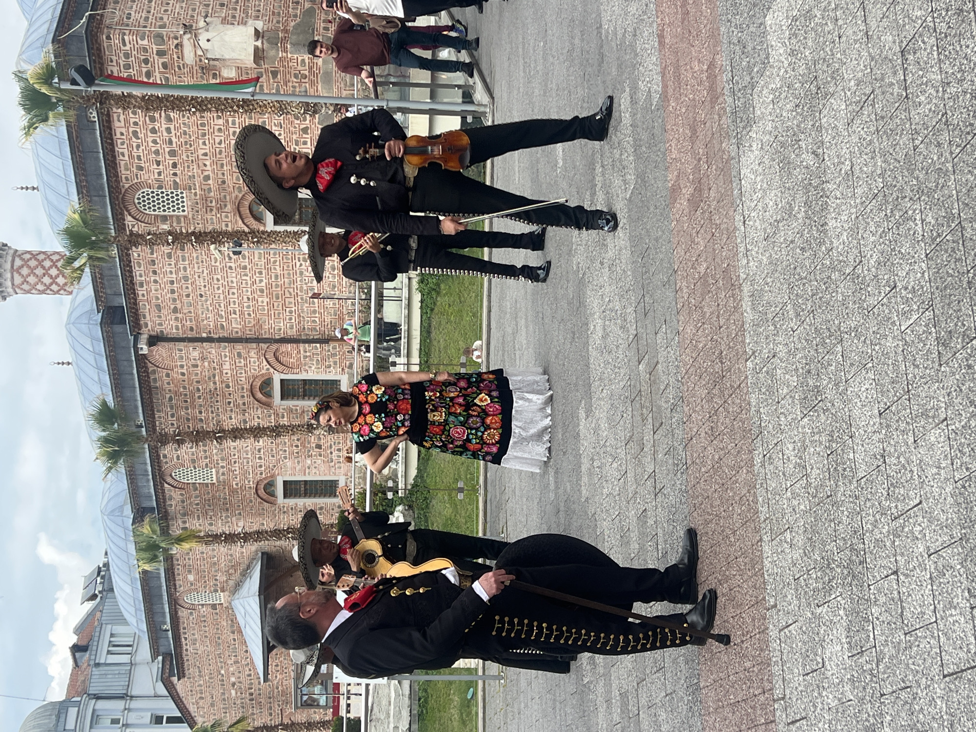 In Plovdiv, Bulgaria, a mariachi band stands in front of the Dzhumaya Mosque, dressed in traditional black attire with silver accents, sombreros, and red bow ties. One member holds a violin and bow; another carries a guitar, while a woman in a colorful, embroidered dress with a white lace hem stands among them. A grey stone-paved square surrounds the scene, with other pedestrians visible in the background.
