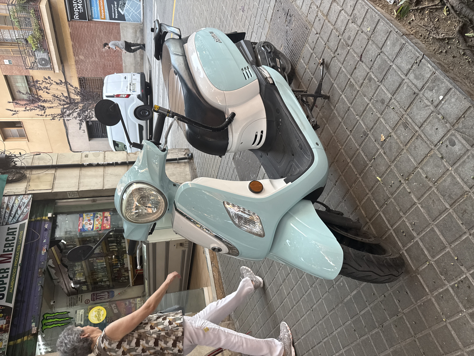 In Barcelona, Spain, a light blue and white scooter is parked on a tiled sidewalk in front of a store. The store has a large glass window displaying various products and a sign that reads "SUPER MERCAT". An older woman with grey hair, wearing a patterned sleeveless top and white pants, walks away from the scooter and towards the store. In the background, a white van is parked on the street, and a man in a grey t-shirt and dark pants walks further down the street. Buildings with windows and balconies are visible in the background. The sidewalk is made of square gray tiles with some debris on it. The lighting suggests it is daytime.