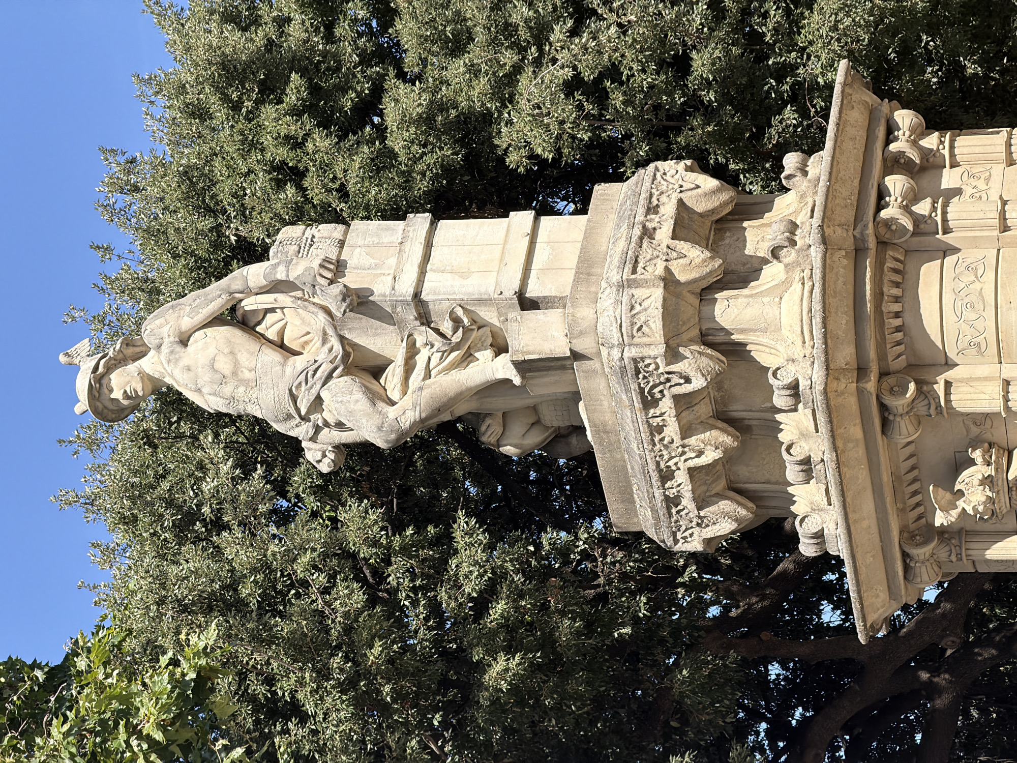The image shows a full-length stone statue of a male figure, likely a mythological or historical character, on a multi-tiered stone pedestal. The statue is partially draped, with muscular anatomical detail visible. The figure wears a winged helmet and appears to be leaning on a column or block. The pedestal is ornately carved with decorative elements. Behind the statue, a dense canopy of green trees forms a backdrop against a clear blue sky. The statue and pedestal are made of light-colored stone, possibly limestone or sandstone, and show signs of age and weathering. The overall impression is of an outdoor public monument. This monument is located in the Parc de la Ciutadella in Barcelona, Spain.