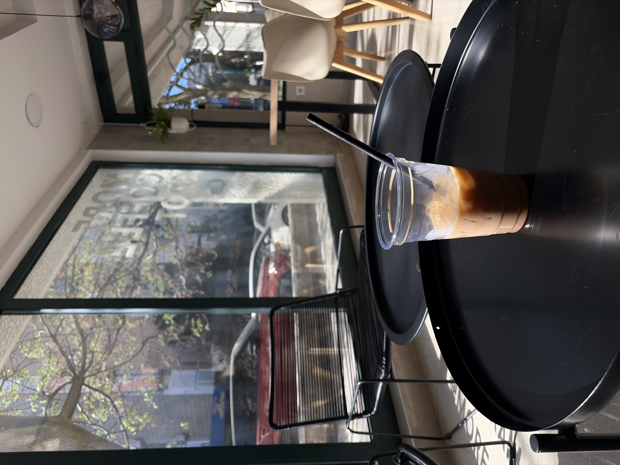 The image displays an indoor scene, likely a cafe or restaurant, on a bright, sunny day in Budva, Montenegro. The main subject in the foreground is a clear plastic cup containing iced coffee with ice cubes and a black straw, placed on a round, black table. The coffee appears light brown and creamy.
The interior features modern furnishings, including additional black round tables and chairs with black metal frames, as well as chairs with light-colored seats and wooden legs visible in the background. A small potted plant hangs from the ceiling.
Through a large window, a vibrant tree with green leaves is prominently visible, suggesting spring or early summer. Reflections on the window show parts of urban buildings and a red car, indicating a street view. The strong sunlight casts shadows, confirming it is daytime with clear weather. Visible text on the window, possibly a decal or reflection, clearly shows the letters "OO" as part of a larger word, likely "COFFEE," reinforcing the cafe setting. No people are present, conveying a peaceful and quiet atmosphere.