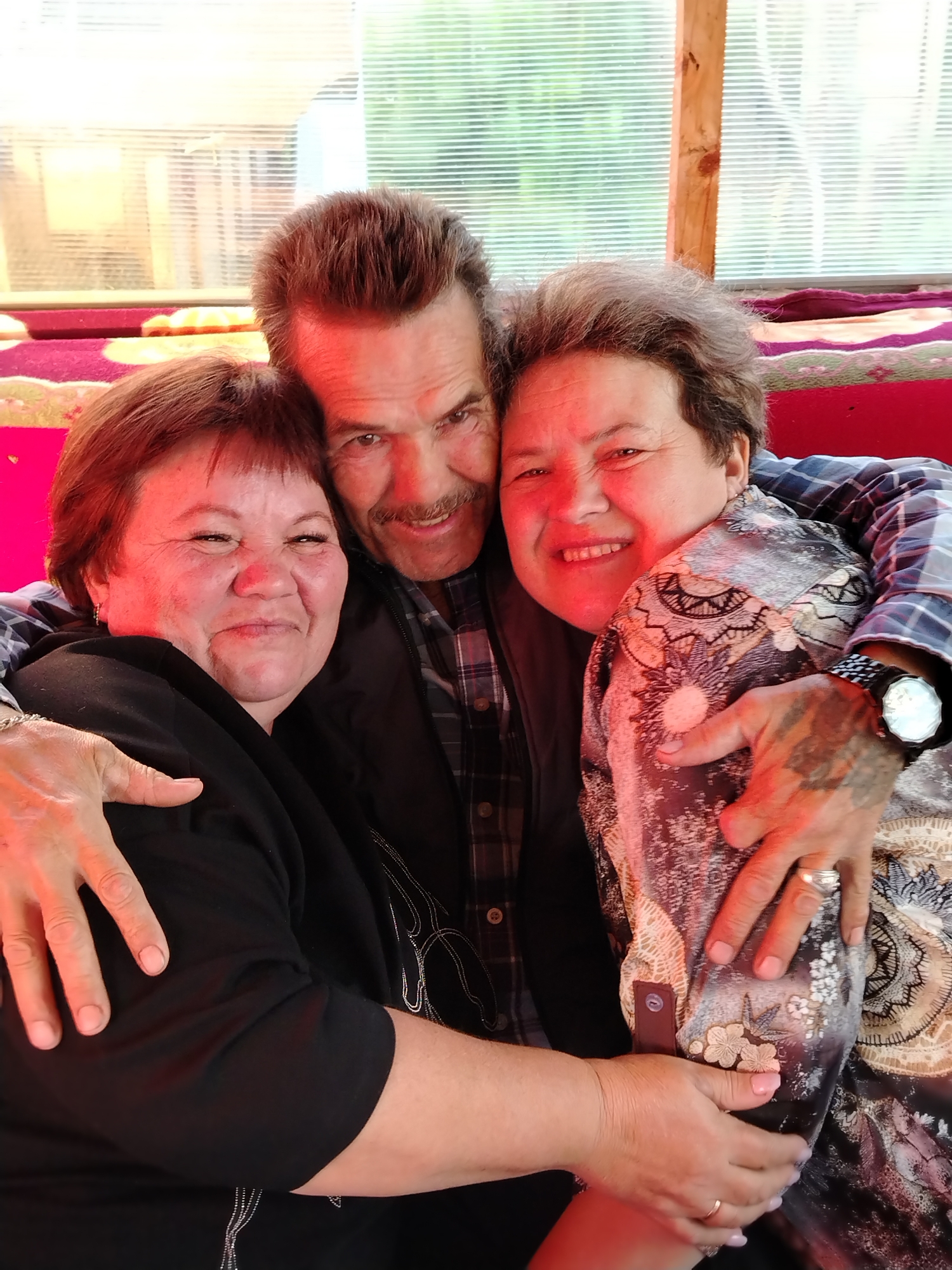 A photo depicts three people embracing in what appears to be an indoor setting. A man with dark hair, a mustache, and a plaid shirt is positioned in the center, smiling. He has his arms around the two women on either side of him. The woman on the left has short, dark hair and is wearing a black top. The woman on the right has short, dark hair and is wearing a patterned dress. Her right hand and wrist have a tattoo and she is wearing a wristwatch and ring. They all appear to be smiling. The background shows a window, part of a red seat cushion and a wall with metal or translucent sheets. The location is in Гожан, Russia.