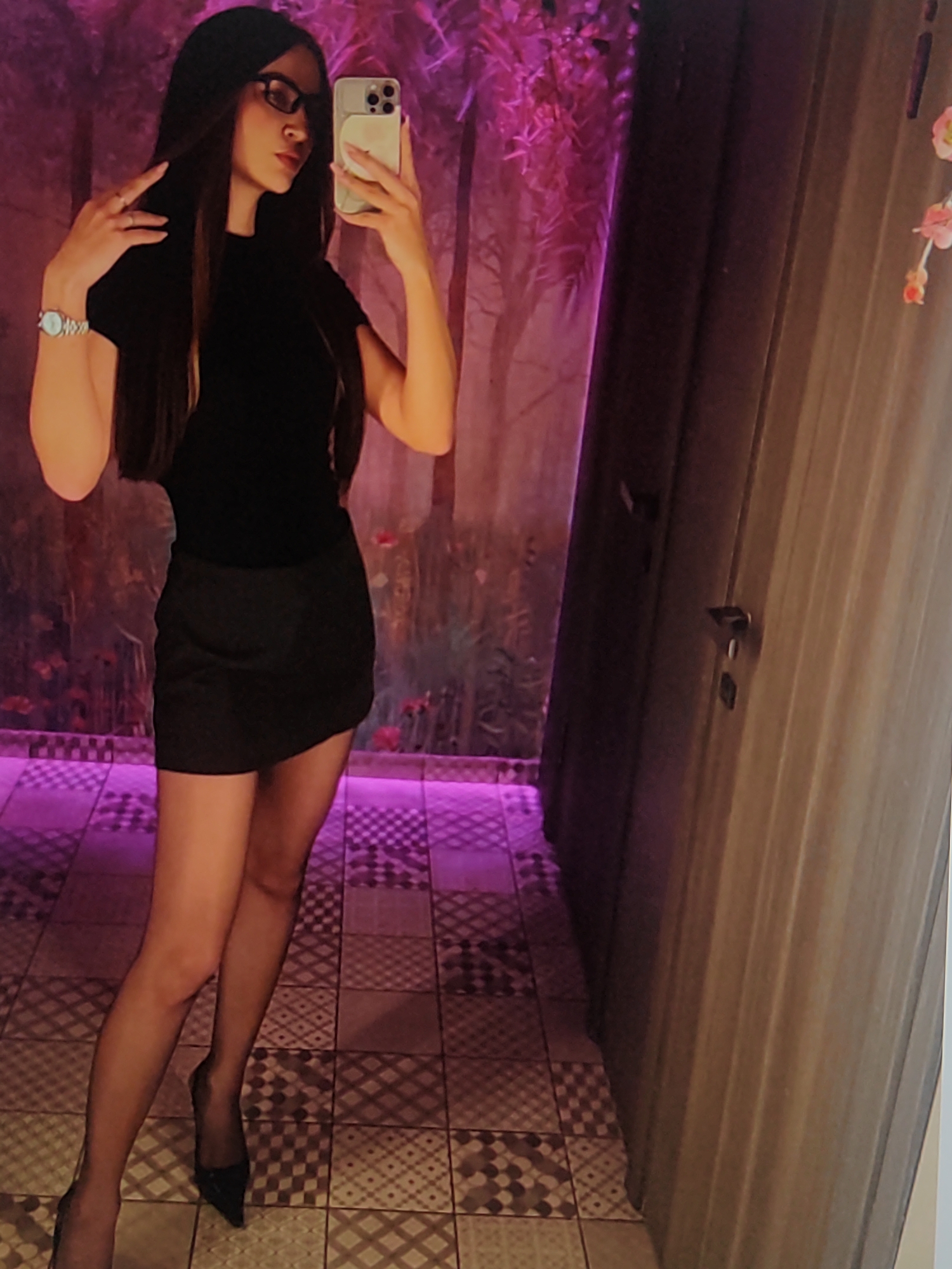 A young woman is taking a mirror selfie in a room with purple lighting. She has long, dark hair and is wearing a black, sleeveless top, a short black skirt, sheer black stockings, and black pointed-toe heels. She is also wearing glasses and a watch on her left wrist. The background appears to be a wall with a floral pattern or mural, illuminated by purple lights that cast shadows. To her right, there's a wooden door with a handle. The floor is tiled with a patterned design. The overall ambiance suggests an indoor setting, possibly a dressing room or a creatively lit space in Fergana City, Uzbekistan. The woman's pose and expression are confident and poised, as she holds her phone up to capture the reflection.