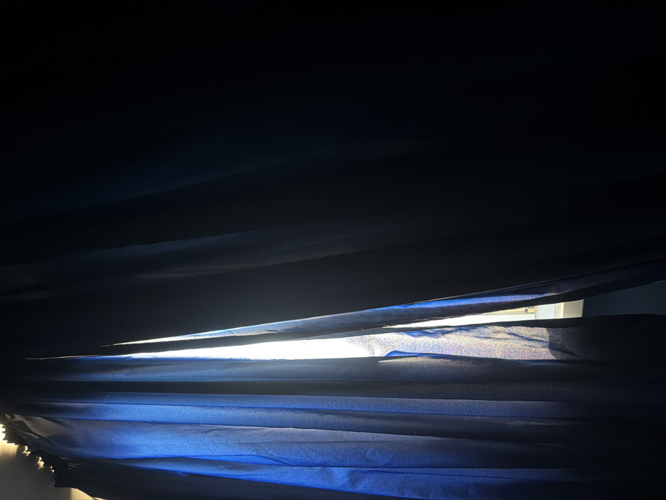 The scene is an interior view, located in Sofia, Bulgaria. The image depicts two dark blue or black curtain panels, likely blackout material, hanging across a window. The curtains are partially open, creating a vertical, luminous gap approximately one-fifth of the image width from the left edge. A strong beam of natural light emanates from this gap, illuminating the inner edges of both curtain panels and rendering the left panel a distinct blue hue. The rest of the room is obscured in deep shadow, indicating minimal light penetration beyond the gap. The bottom of the light gap reveals a faint outline of a window frame or sill. No individuals, additional subjects, specific objects, or external city landmarks are visible. The interaction is solely between the incoming light and the curtains, creating a high-contrast environment.