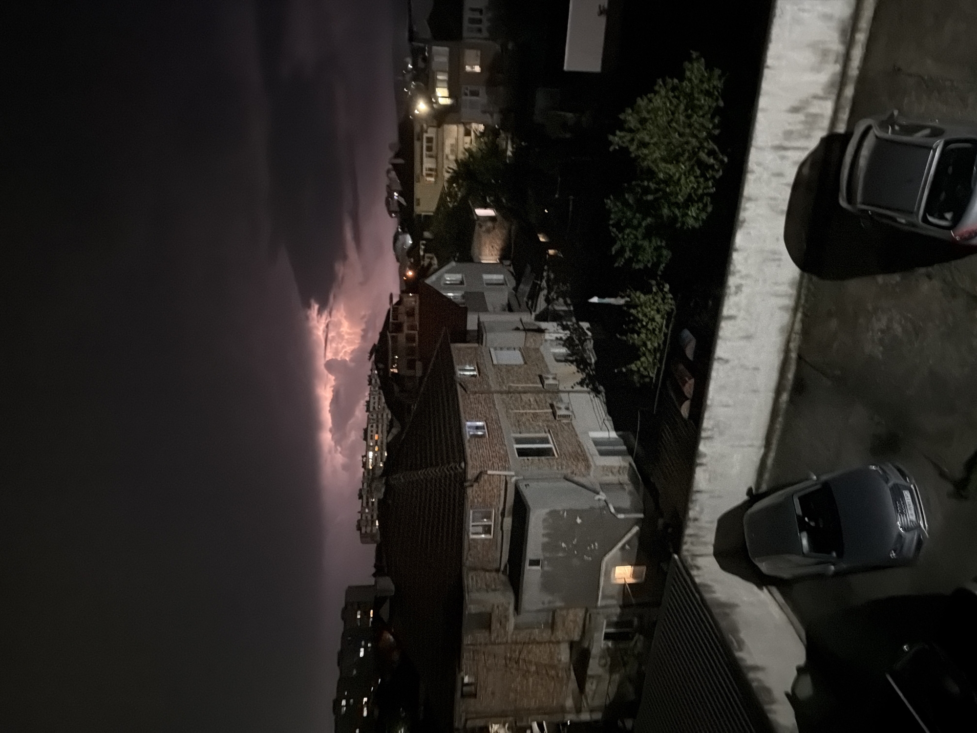 The image shows a nighttime view from a high vantage point, likely a balcony or window, overlooking a city neighborhood. Dark storm clouds are visible in the sky, with a faint glow suggesting either lightning or the setting sun obscured by cloud cover. Below, a dense arrangement of buildings, mostly low- to mid-rise residential structures with red-tiled roofs, are illuminated by scattered streetlights and the faint glow from some windows. Cars are parked on the streets or in designated parking areas. The location is likely in Burgas, Bulgaria given the user input, but this cannot be conclusively determined from the image alone. The scene appears calm despite the impending storm. No people are directly visible in the image.