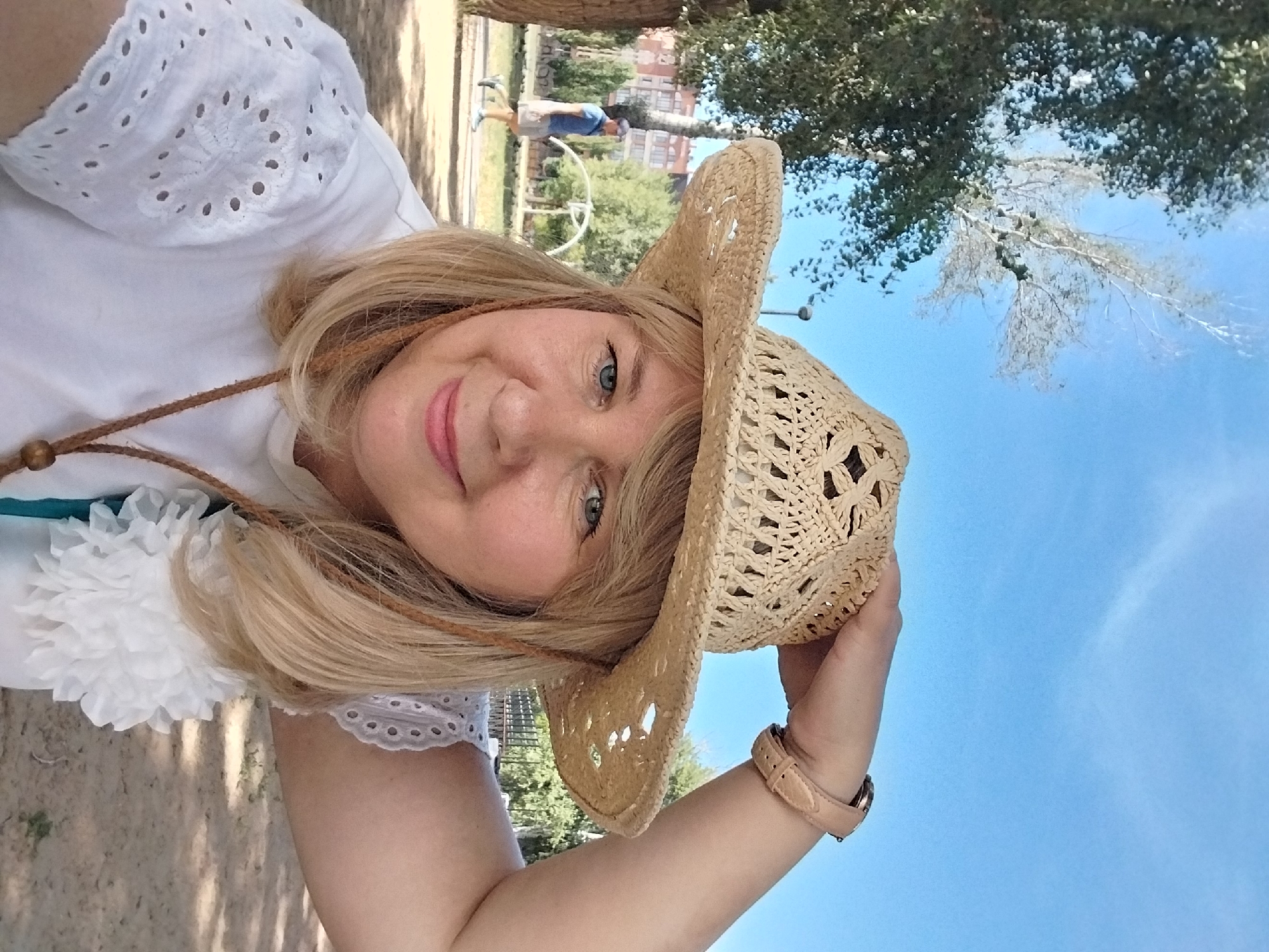 A woman with blonde hair and blue eyes is captured in a selfie. She is wearing a straw cowboy hat and a white shirt with eyelet detailing on the sleeves. A braided cord necklace with a wooden bead hangs around her neck, and a white fabric flower adorns her shirt. She has a watch with a light brown band on her right wrist. Her left hand is holding the brim of her hat.
The background suggests an outdoor setting, possibly a park or public space. Sunlight is filtering through trees, casting shadows on the sandy ground. In the middle ground, a person wearing a blue shirt and light-colored shorts is walking away from the camera. Behind them, there are some trees and a glimpse of buildings in the distance. The sky above is a clear blue.
The image is taken during the daytime, indicated by the bright lighting and shadows. The overall atmosphere appears casual and relaxed. The location is specified as a city park in Orenburg, Russia.