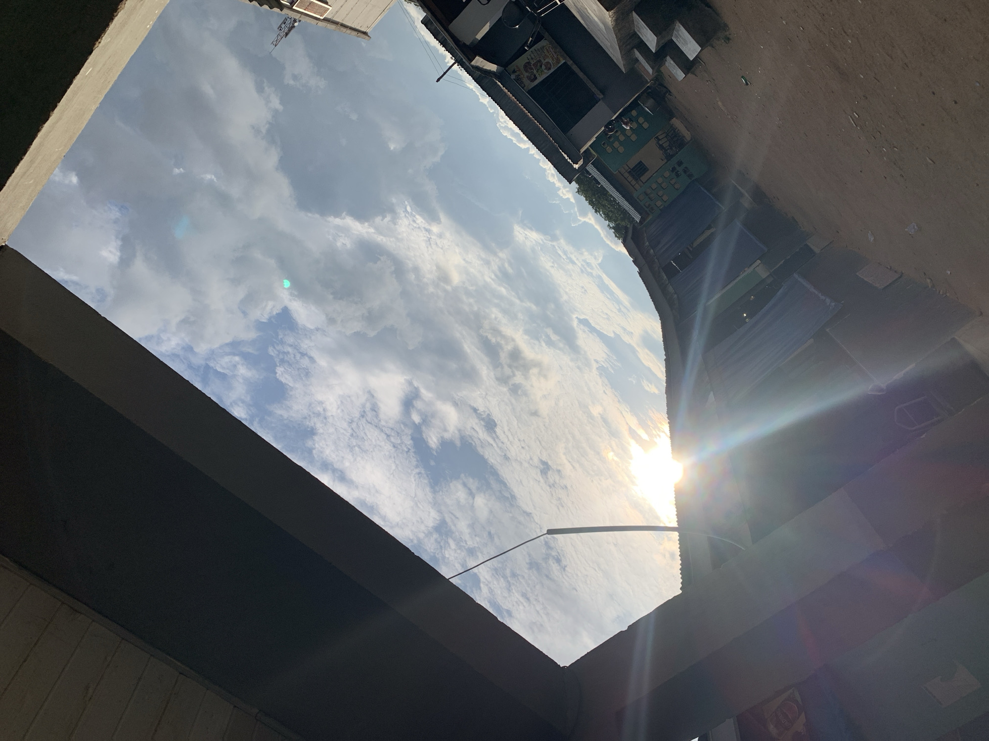 This image, captured at a tilted angle, depicts an outdoor scene in New Stadium, Nigeria, viewed looking upwards towards a partly cloudy sky at either sunrise or sunset. The sun, a brilliant luminous disc, is positioned in the lower right portion of the sky, emitting strong lens flare and distinct sun rays. The sky features a mix of white and grey stratocumulus clouds against a clear blue background.
The upper frame is dominated by dark, angular architectural elements. A large, dark grey concrete structure, possibly the underside of an overpass or a substantial beam, extends diagonally across the top left towards the mid-right, presenting a textured surface on its visible edge. Another dark, undefined structural element frames the top right corner.
A row of single-story commercial or residential buildings with corrugated metal roofs runs diagonally from the lower left towards the mid-right. These structures display varied exterior finishes, including sections painted green and grey. Visible details on the buildings include rectangular windows, dark recessed doorways, and faded signage; one sign on the rightmost building clearly displays text and graphics, possibly a menu or advertisement. On the far right, a set of concrete block steps ascends towards one of the structures. The ground in the immediate foreground is unpaved, composed of reddish-brown dirt or sand. A thin, dark pole or wire extends from the left framing structure into the bright sky, angled towards the sun. Sparse green foliage is discernible on a distant elevated terrain behind the buildings on the right. No individuals, explicit actions, or direct interactions are visible within the frame.