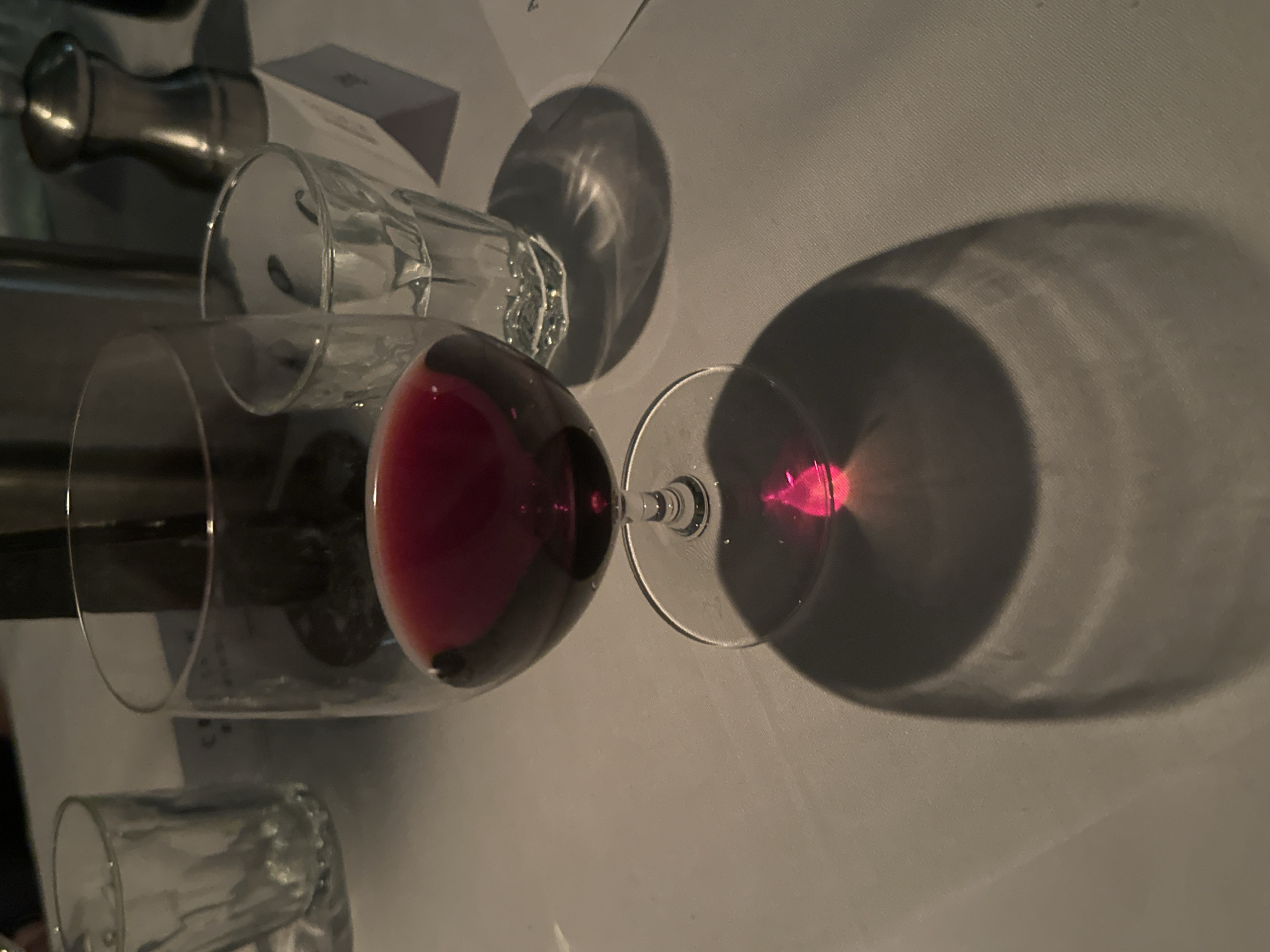 The image, taken in London, United Kingdom, presents a close-up view of a dining table setting under dim, artificial lighting. The central focus is a wine glass holding a quantity of deep red wine. To its left, another empty, clear glass is positioned, possibly a water glass or another type of drinking vessel. In the background, a metallic object, which appears to be part of a wine accessory like a stopper or pourer, and a white card or paper (potentially a menu or place card) are visible.
The low lighting creates prominent, elongated shadows of the glasses on the light-colored tablecloth. A notable detail is the distinct refraction of light through the red wine, casting a vivid pinkish-red spot within the shadow of the wine glass, adding a splash of intense color to the otherwise subdued palette. The tablecloth exhibits a subtle texture.
The scene suggests an indoor setting, likely a restaurant or a formal dining environment, indicative of an evening meal or social gathering. No people are present, and no specific activity is captured beyond the static arrangement of the objects. A faint numeral "1" can be seen on the white card in the background, but no other discernible text is visible. The overall mood is quiet and intimate due to the ambient light conditions.