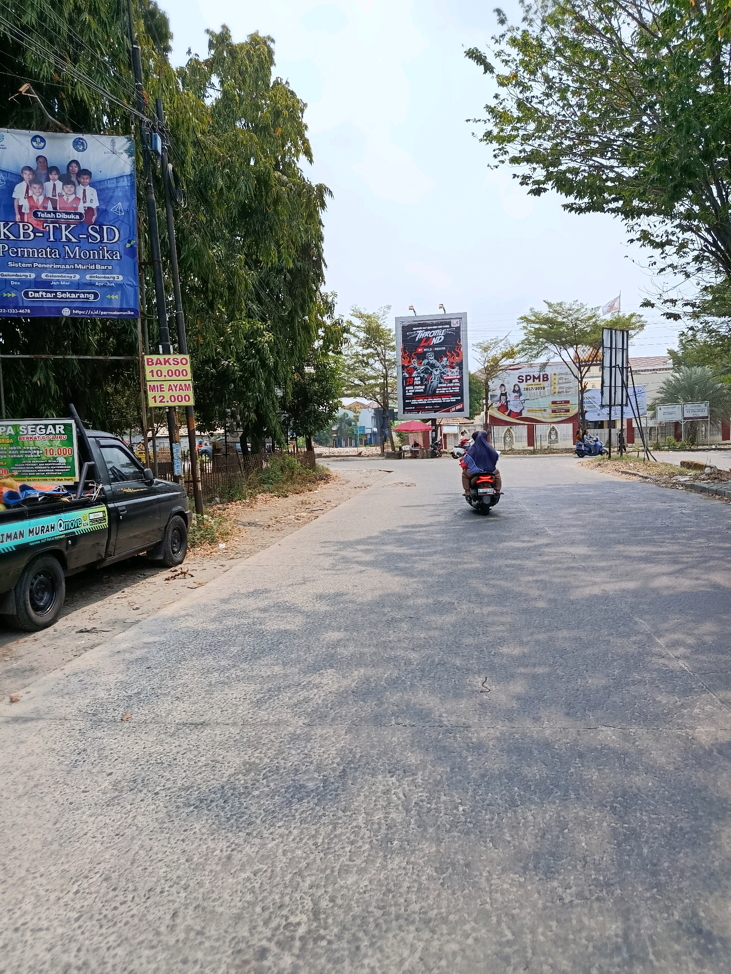 The image captures a street scene in Bekasi, Indonesia, on a bright, sunny day with a clear sky. The road is paved and appears to be in moderate condition, with some cracks visible. Lush green trees line the sides of the street, casting shadows on the asphalt. Several billboards are prominently displayed along the roadside, advertising educational institutions and events.
On the left side of the road, a black pickup truck is parked, displaying advertising banners for "ESPA Segar" and "OTTIMO MURAH". A motorcycle is parked behind the pickup. Further down the street, a red umbrella shades a small food stall.
In the middle of the road, a person wearing a blue headscarf and dark clothing rides a scooter, moving away from the camera. Another person on a scooter, with blue clothing and a white head covering, is visible further ahead in the distance, also moving away. Several other individuals are scattered along the sidewalk and in front of buildings.
Dominant billboards include one for "KB-TK-SD Permata Monika", a school with children pictured and details about enrollment. Another large billboard advertises an event called "THROTTLE LAND". A third billboard promotes "SPMB 2027/2028", likely referring to a school entrance exam. In the background, there are buildings, some with a light-colored facade, and a white flag flying. The overall impression is a busy, everyday scene in an urban environment.
