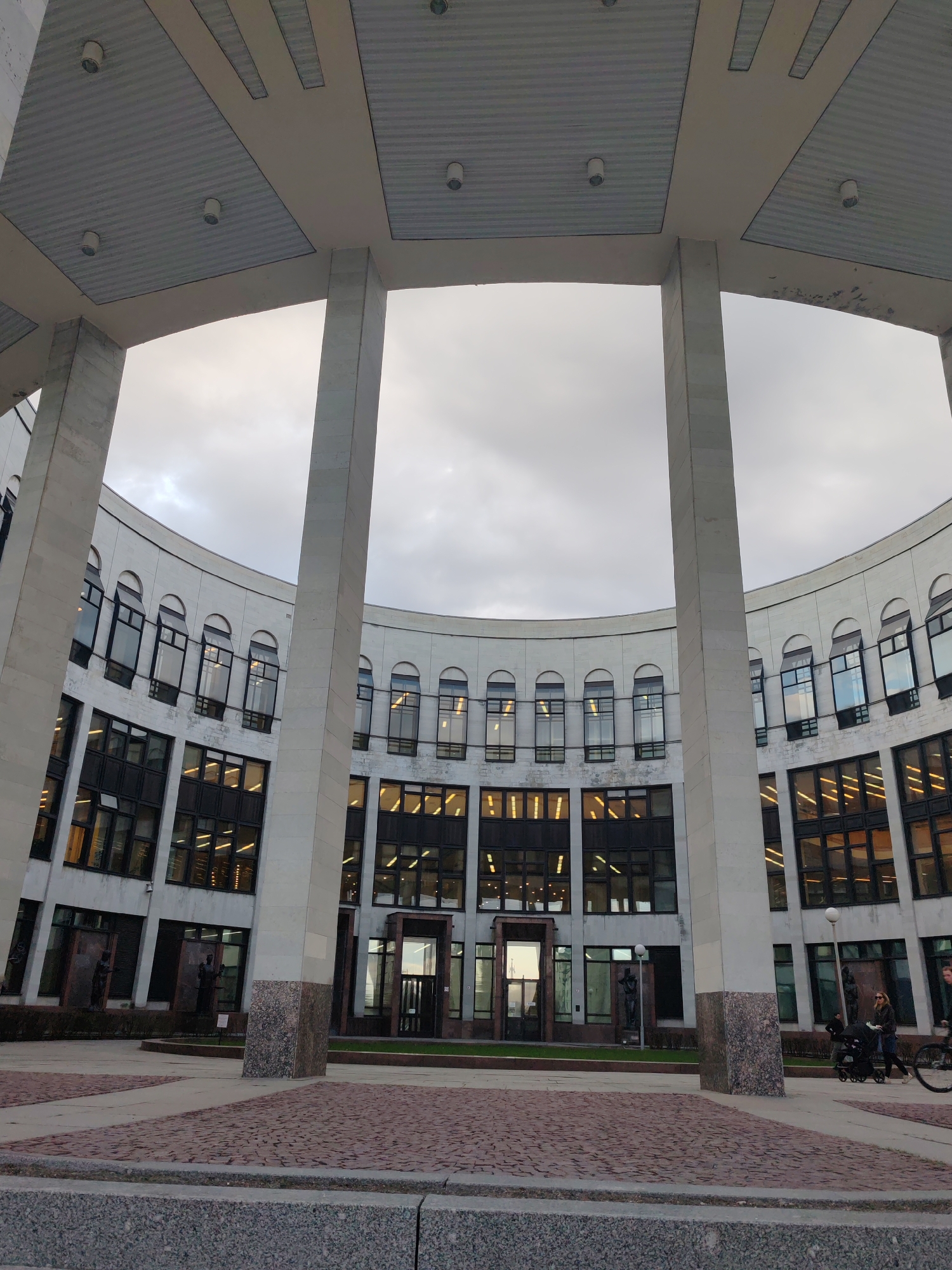 An eye-level shot captures a grand, curved building with modern architecture, viewed from a spacious outdoor plaza in Saint Petersburg, Russia. The building is light-colored, possibly stone or concrete, and features multiple stories. The upper floors have a repeating pattern of dark, rectangular windows topped with rounded arches, while the lower floors display larger, multi-paned rectangular windows. Many interior lights are visible through the windows, suggesting either dusk, night, or an overcast day.
Four massive, light-colored square columns dominate the foreground and midground, supporting a high, flat overhead structure that extends from the building. This ceiling features recessed paneling and several circular light fixtures. The sky beyond the building and columns is uniformly grey and overcast, indicating a cloudy day.
The ground of the plaza is paved with a distinctive pattern of reddish-brown brick-like material, bordered by grey stone. On the far right side of the image, two individuals are visible: one person is pushing a dark-colored baby stroller, and another person is partially seen, appearing to be on a bicycle. These figures suggest a public space with pedestrian traffic. Dark, abstract sculptures or figures stand in niches near ground-level entrances along the building's facade. A lamppost is also visible to the right. No discernible text is visible in the image.